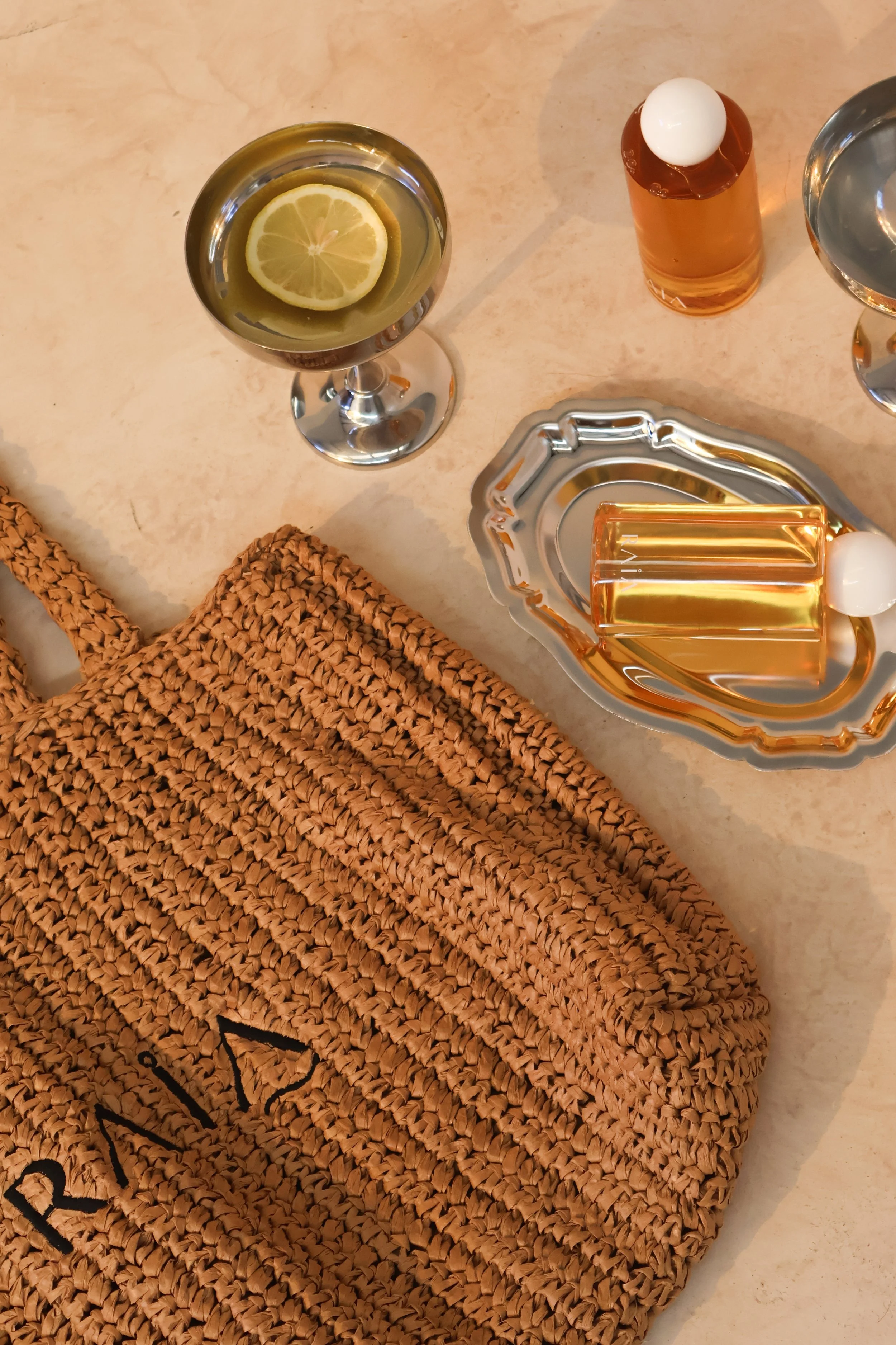 A woven straw handbag, a glass with lemon water, two bottles of skincare products, and a decorative dish with a gold object on a beige marble table.