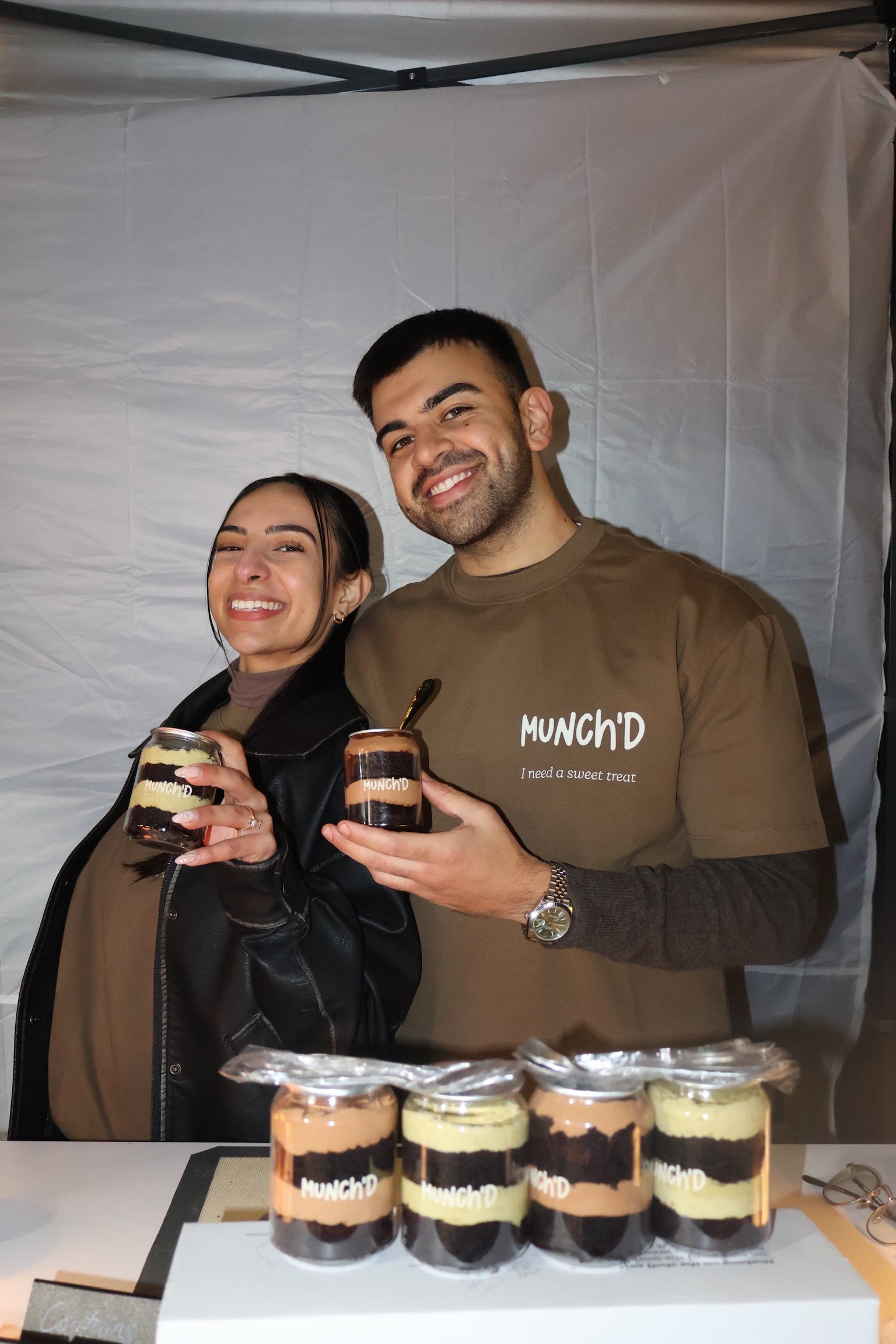 Two smiling people holding jars of layered dessert at a booth with more jars on the table, branded with 'MUNCH'D'.
