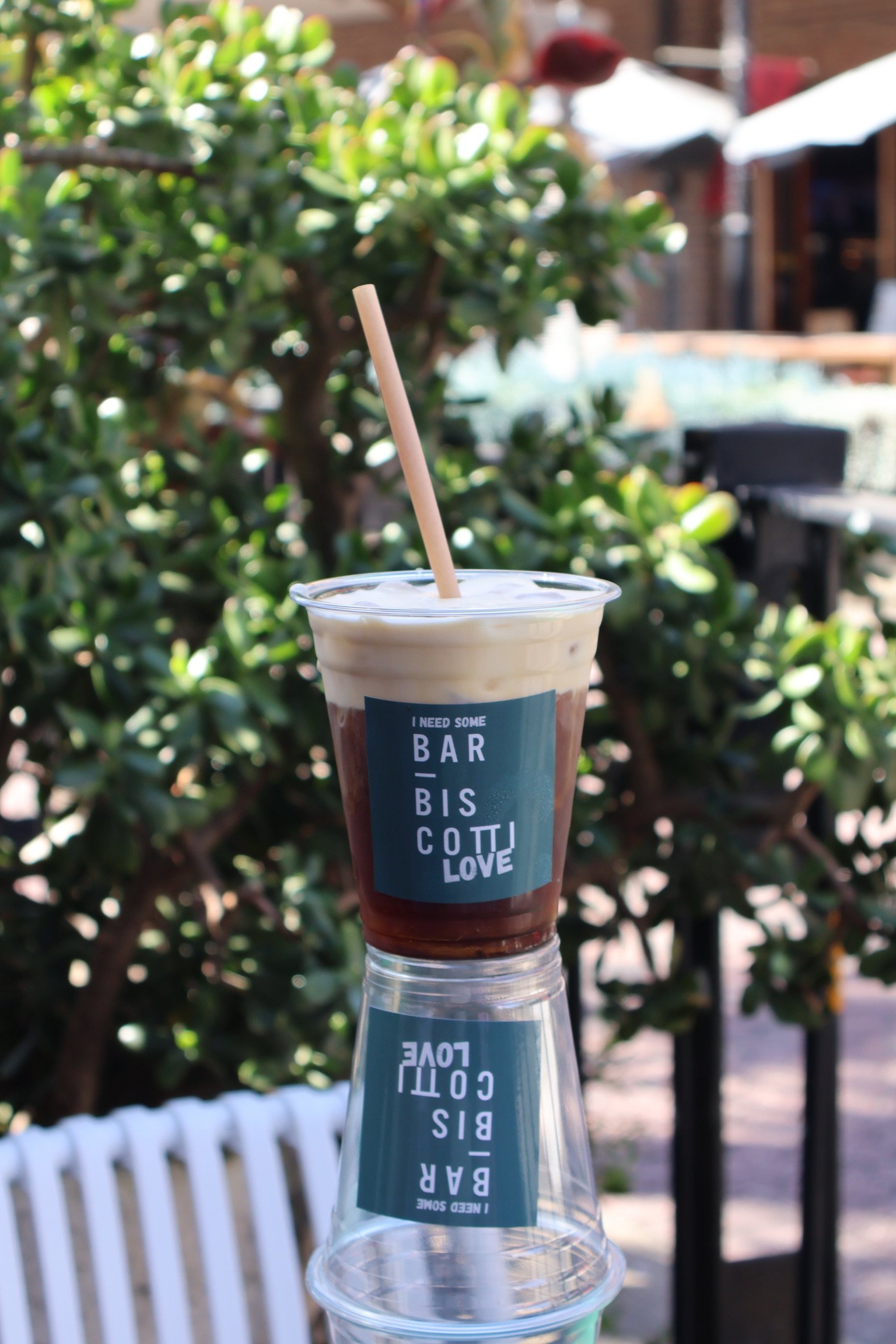 A plastic cup of iced coffee with a straw, placed upside down on top of an upside-down glass, outdoors with greenery and a wooden building in the background.