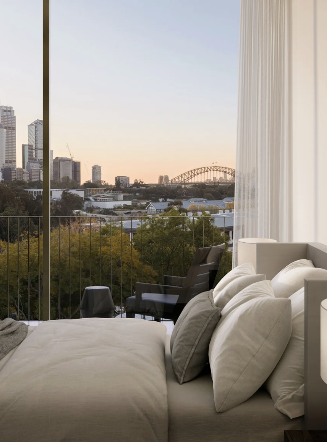 View of a city skyline with tall buildings and a bridge in the distance from a bedroom window at sunset. The room has a bed with white linens and pillows, a black outdoor chair, a small side table, and sheer white curtains.