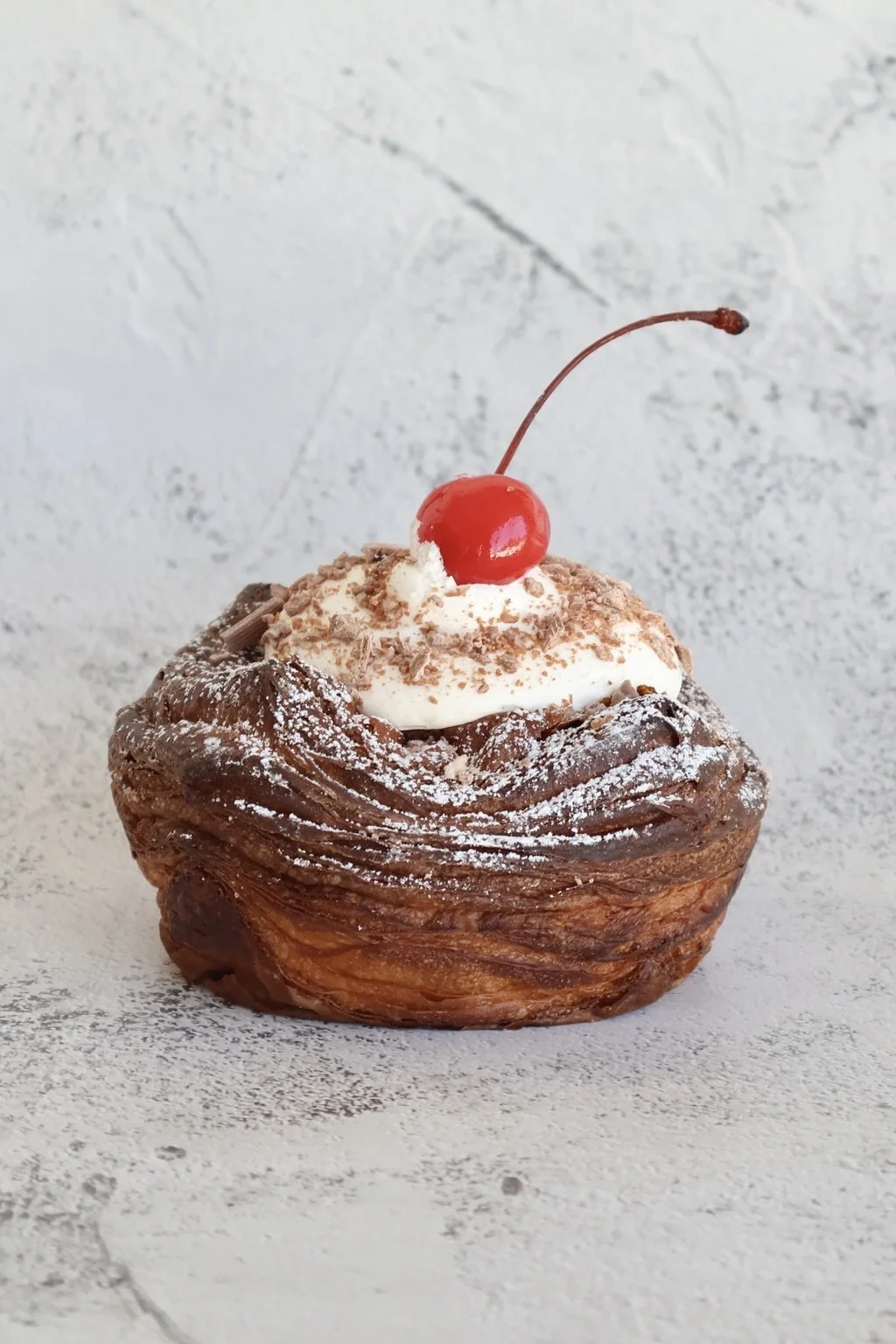 A close-up of a chocolate pastry topped with whipped cream, chocolate shavings, a maraschino cherry, and dusted with powdered sugar.