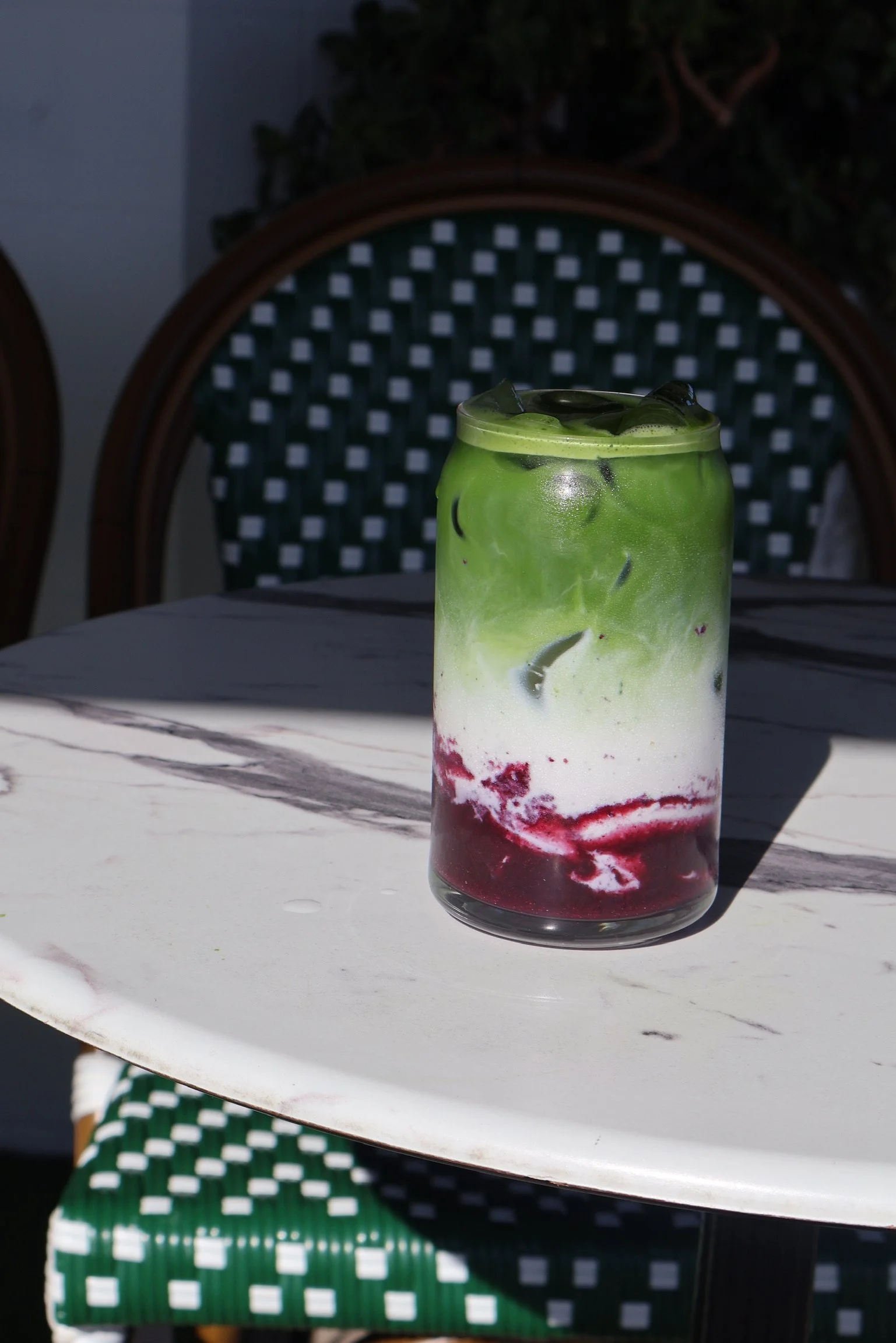 A tall glass of layered green, white, and red beverage with ice cubes on a white marble table outside.