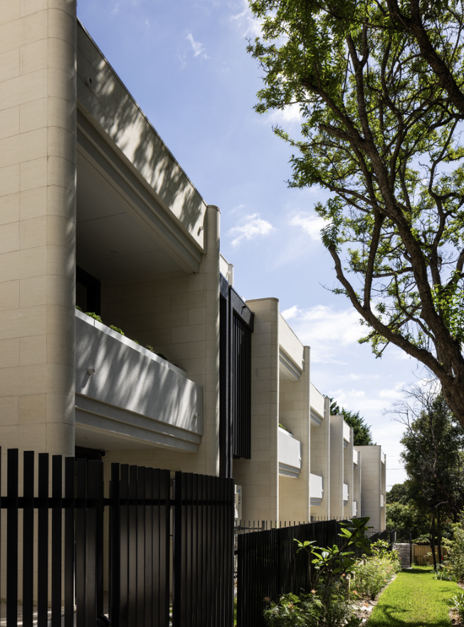 Modern multiple-story white apartment building with balconies, black fencing, green trees, and a bright blue sky.