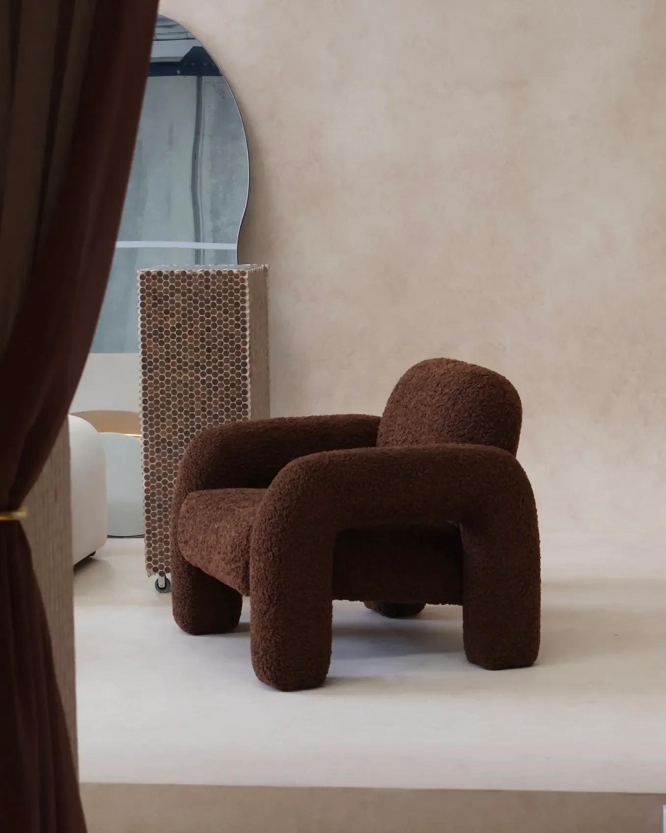 A brown plush armchair with rounded arms and backrest, sitting on a light-colored floor against a beige wall. In the background, a mirror and a decorative panel are visible.