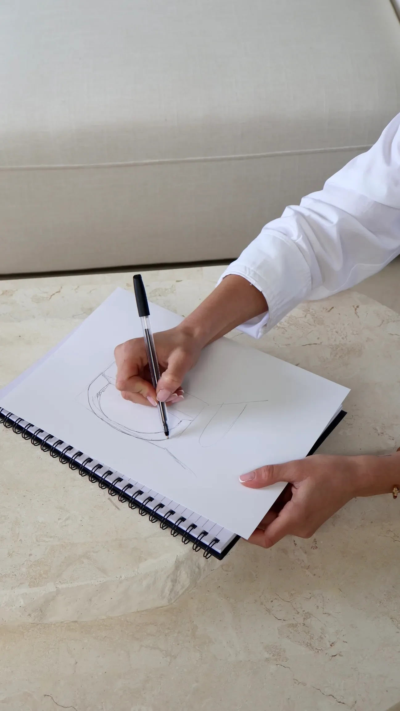 Person drawing on a sketch pad with a pen, wearing a white sleeve shirt, on a marble surface.