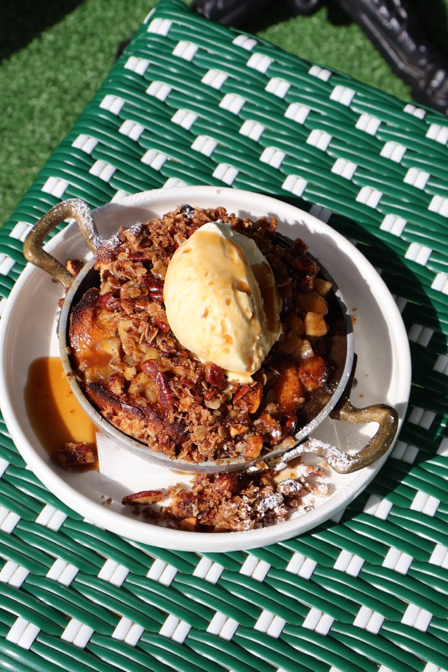 Apple crumble pie with a scoop of vanilla ice cream on top served on a white plate outside on a green and white checkered outdoor table.