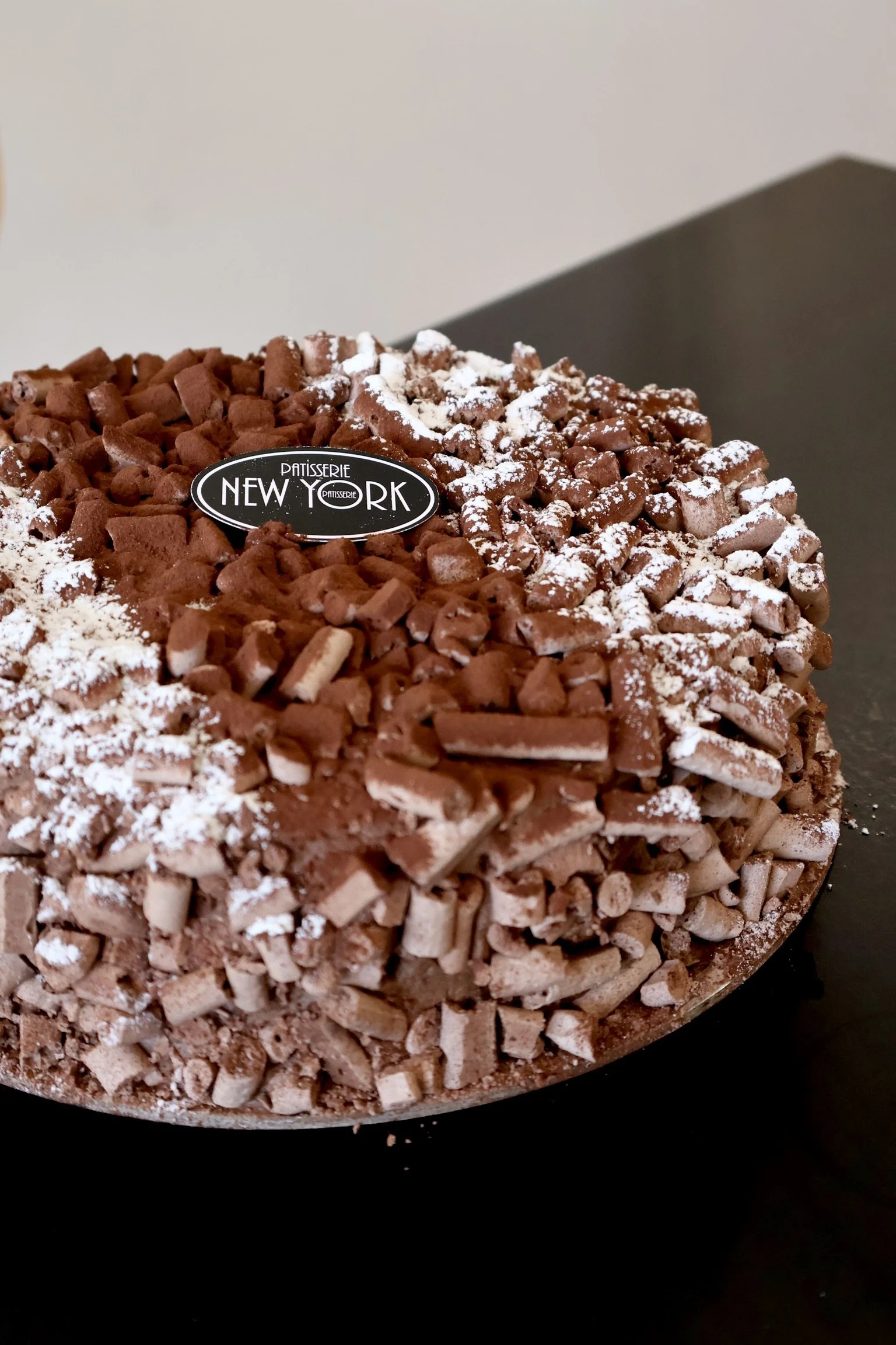 Close-up of a round chocolate cake covered with chocolate shavings and powdered sugar, with a "Patissserie New York" bakery label on top, placed on a dark surface.