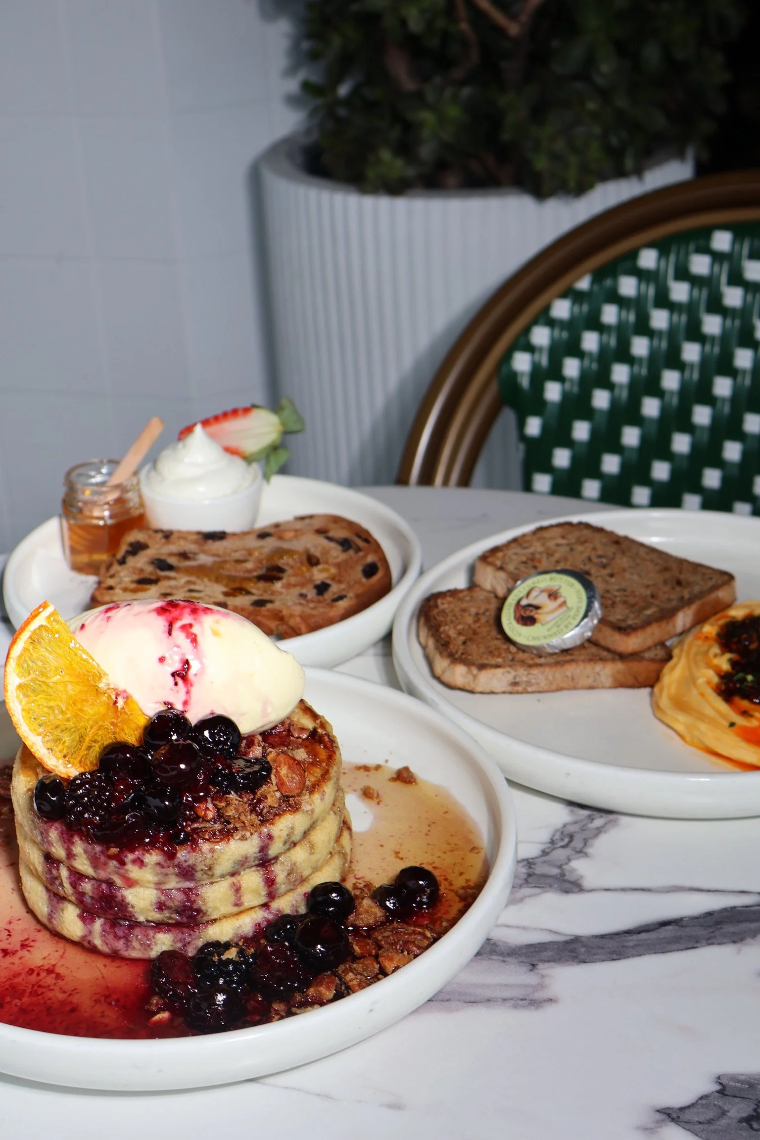 A plate of stacked pancakes topped with berries, ice cream, and an orange slice, served with syrup. In the background, there are plates with strawberries, toast, and breakfast meats on a marble table.