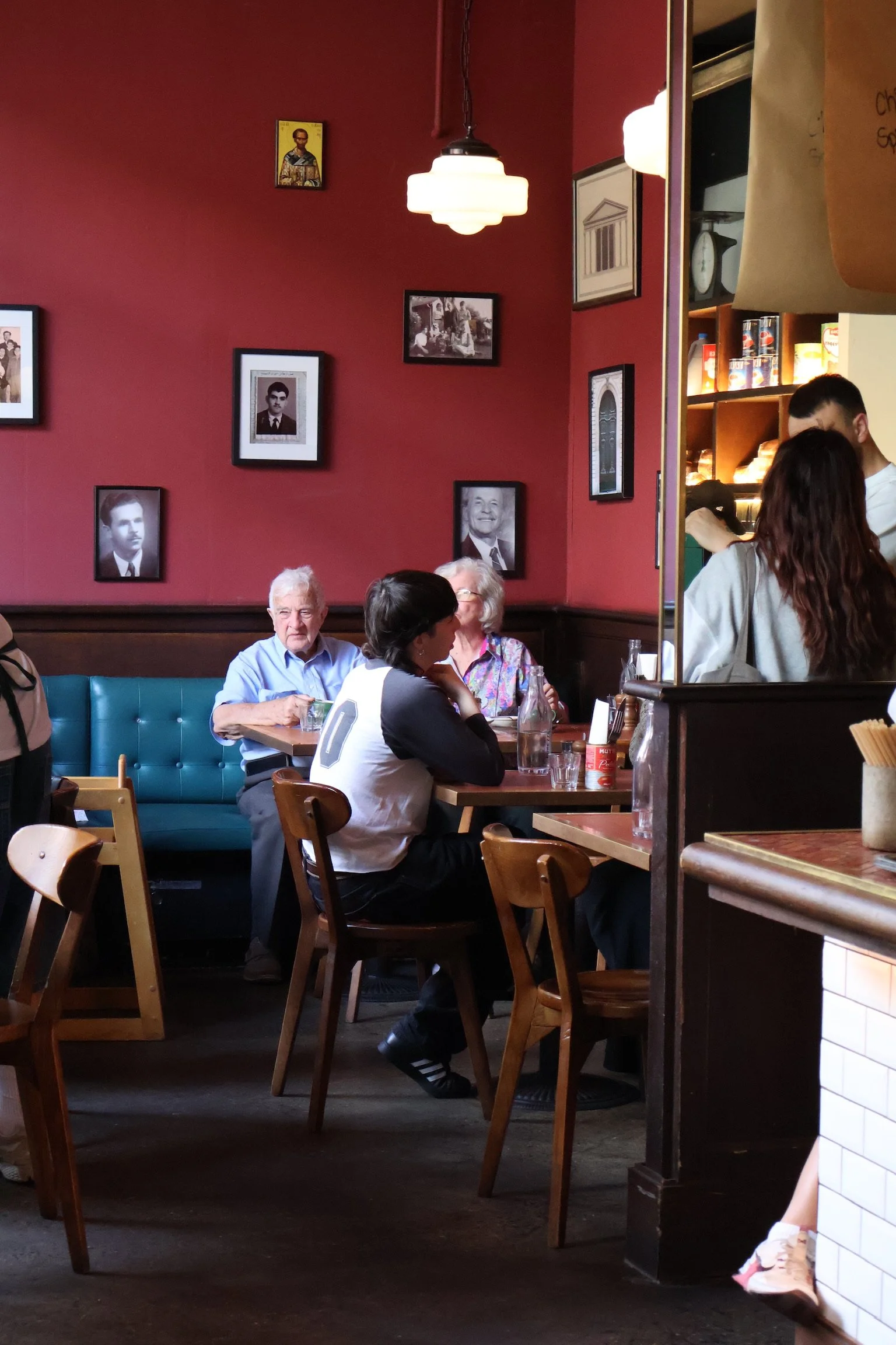People dining inside a restaurant with framed photos on a red wall, a hanging light fixture, and a mirror reflecting part of the room.
