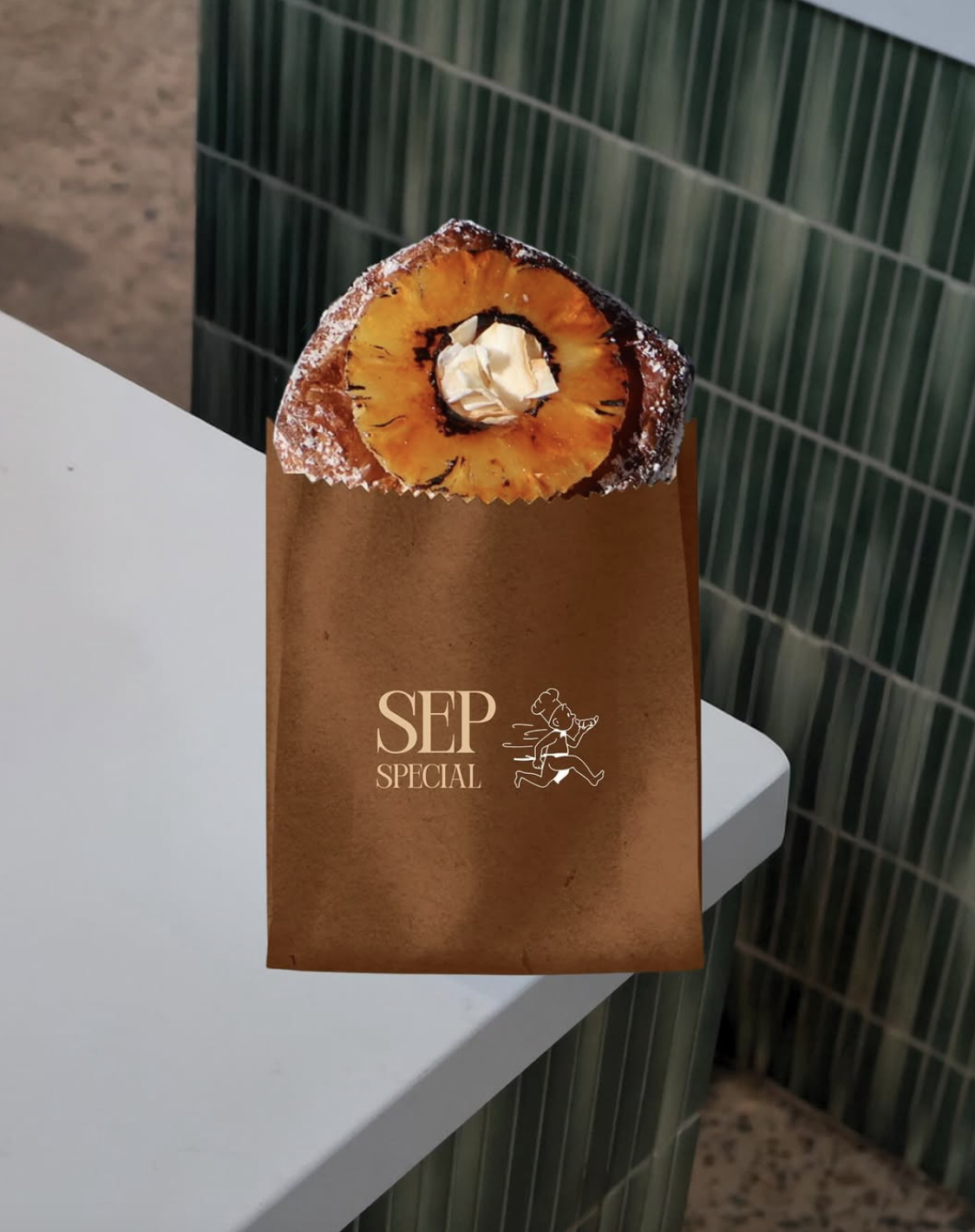 A small pastry topped with a pineapple slice and a dollop of whipped cream, placed on a brown paper bag with 'SEP SPECIAL' and a running chef illustration, on a white surface next to green tiled wall.