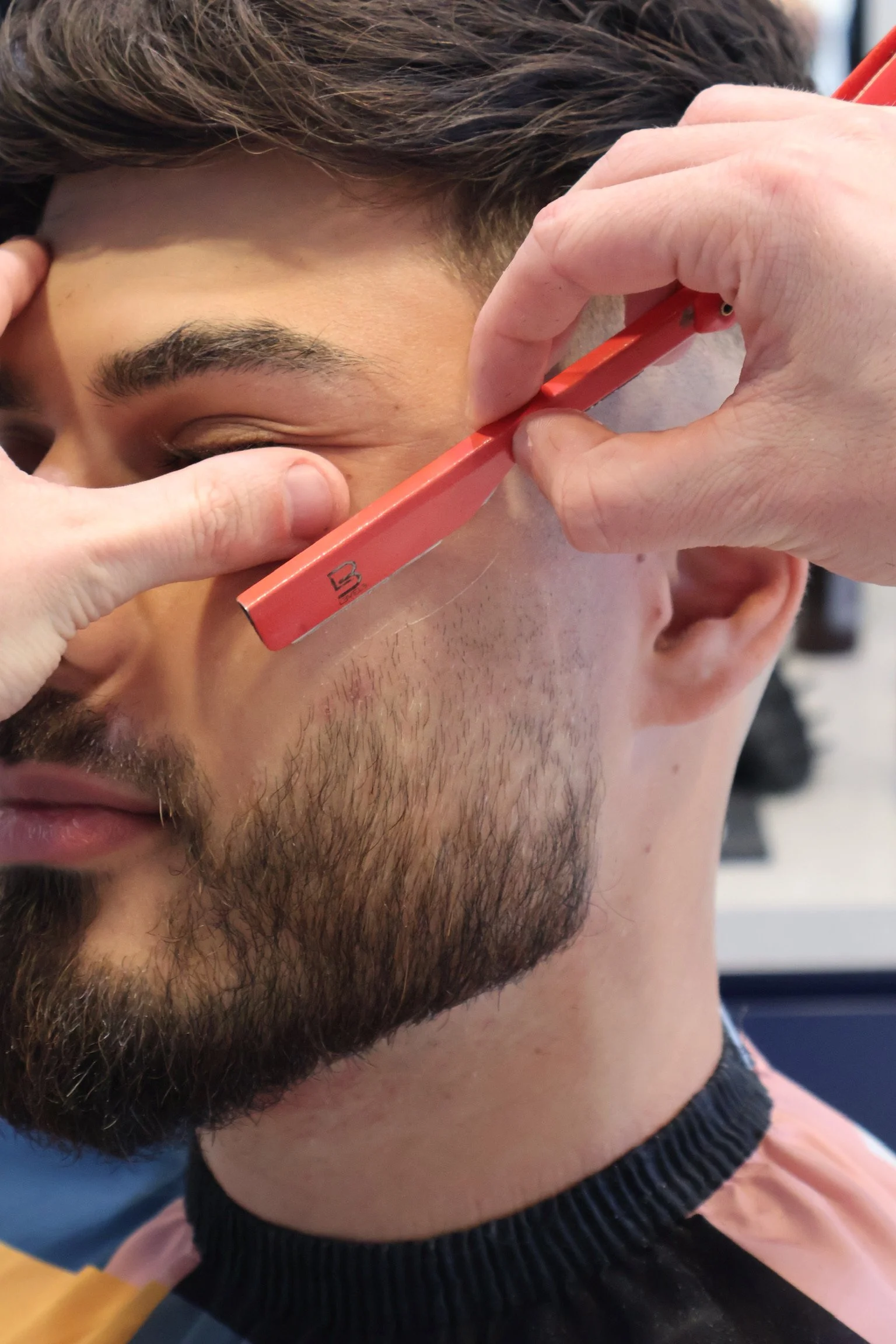 A person getting a facial shave with a straight razor held by another hand, with the person's eyes closed.