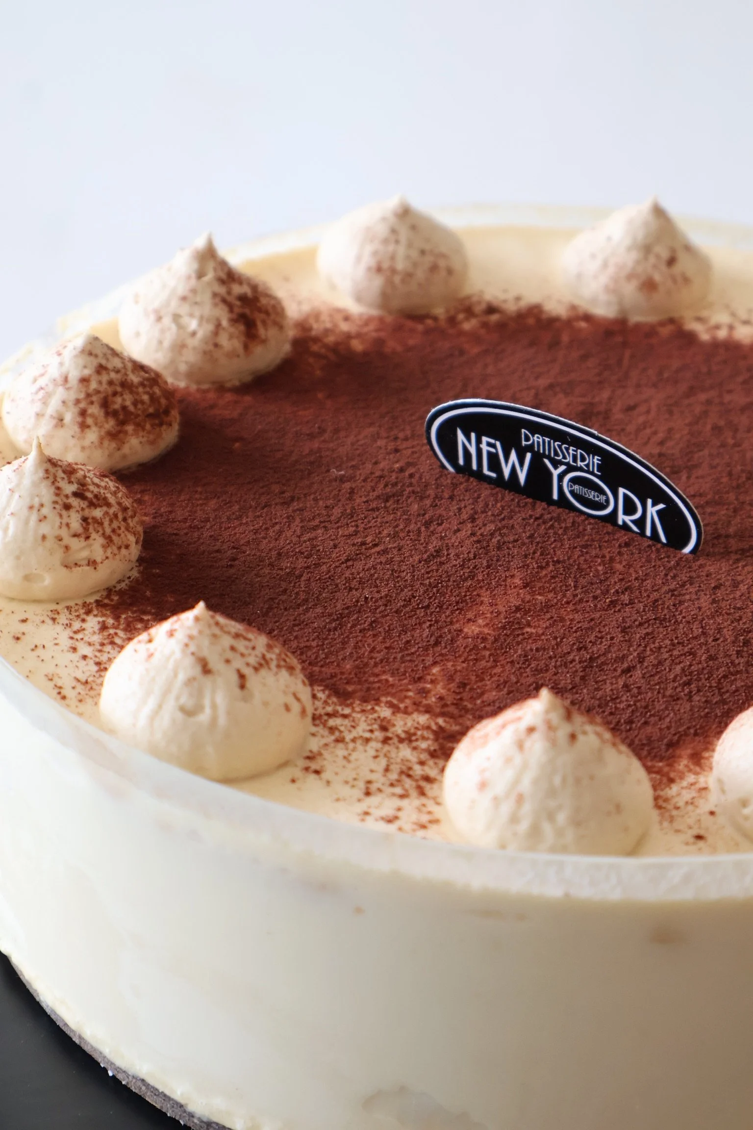 A close-up of a round tiramisu dessert with cream topping and cocoa powder dusted on top, with a black label reading ‘New York’ from Patisserie, and topped with dollops of whipped cream.
