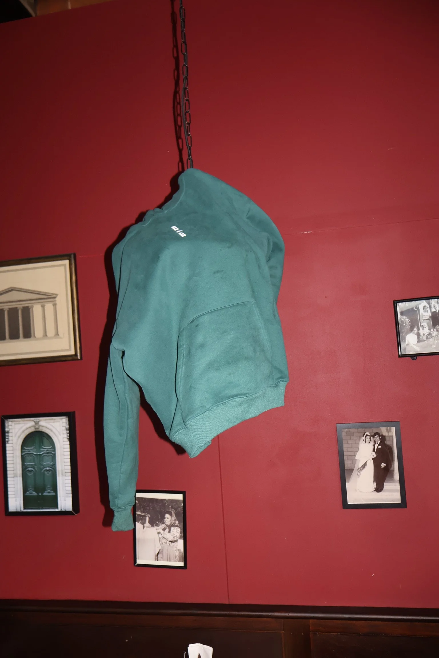 A teal hoodie hanging on a black chain against a red wall, with black and white framed photographs on the wall.