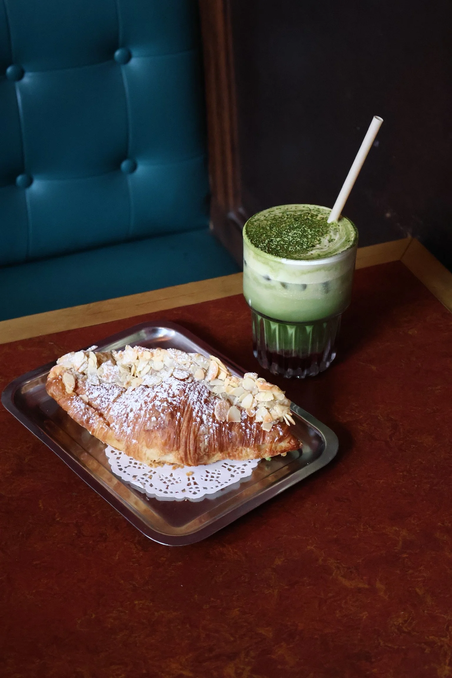 A pastry topped with sliced almonds and powdered sugar on a silver tray next to a green matcha latte with whipped cream and matcha powder on top, on a wooden table in a cafe.
