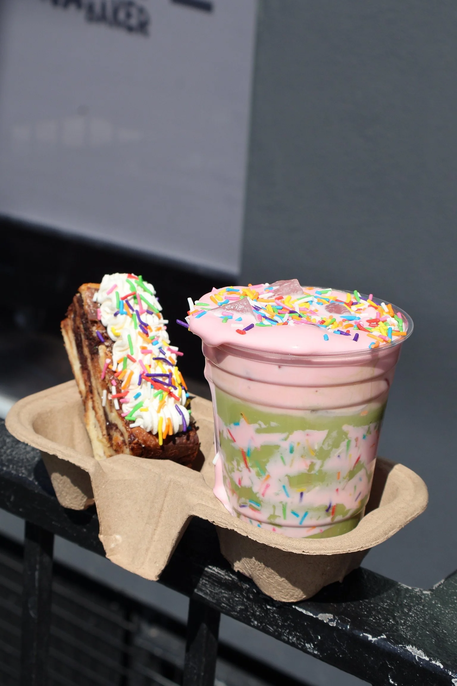 A tray holding a slice of layered cake with white and chocolate layers topped with whipped cream and rainbow sprinkles, and a  colorful ice cream sundae with pink and green layers, pink frosting, and rainbow sprinkles.