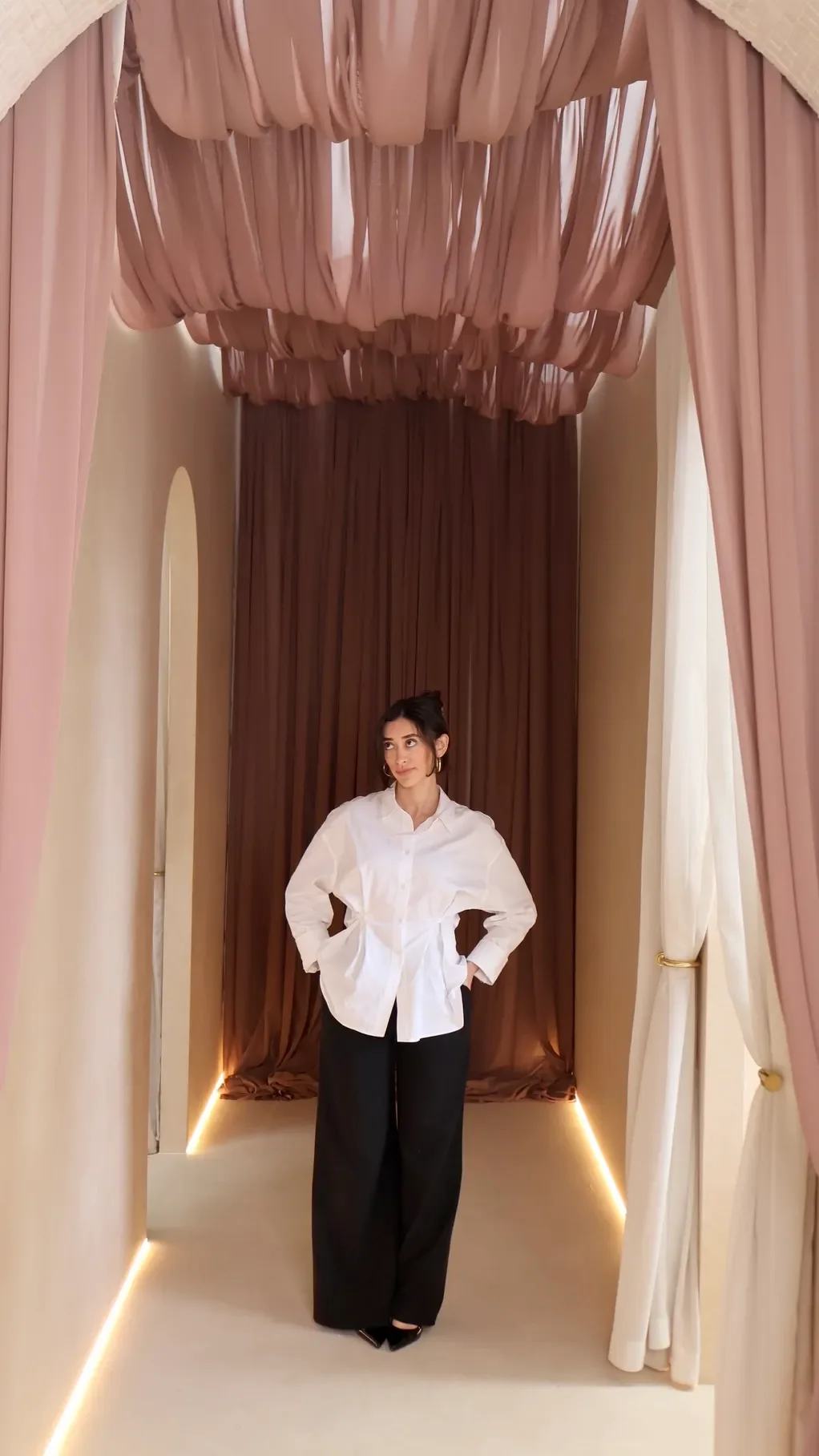 A woman in a white button-up shirt and wide-leg black pants standing in a dressing room with pink and beige curtains.