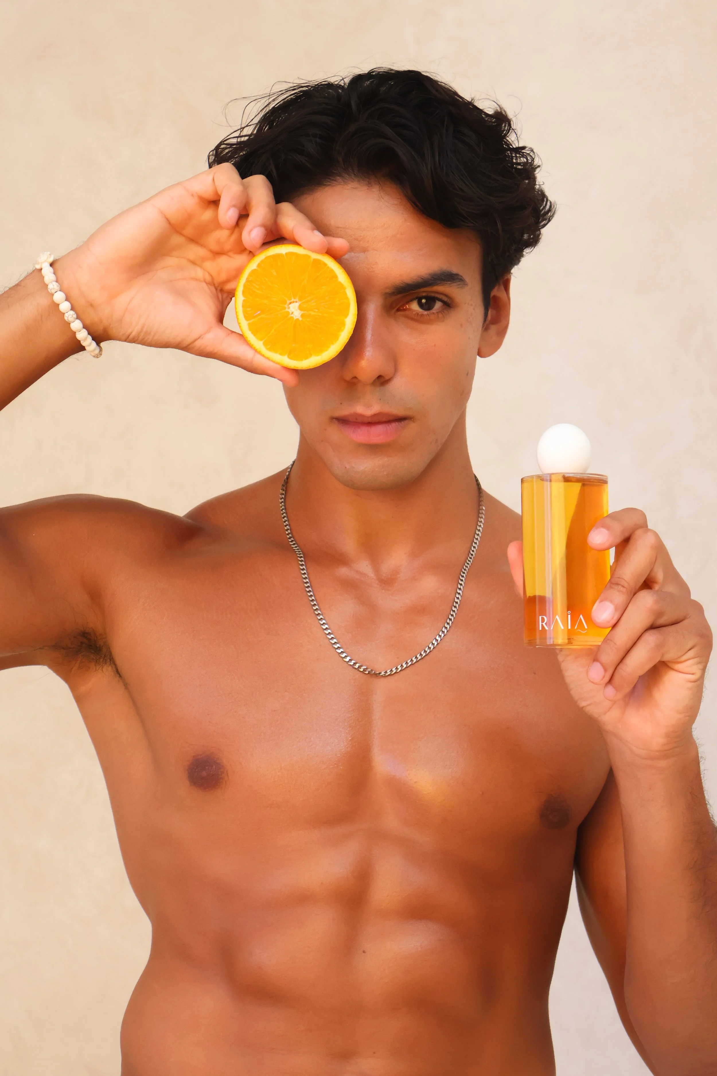 A shirtless young man with dark hair holds a halved orange over one eye and a perfume bottle in his other hand against a neutral background.