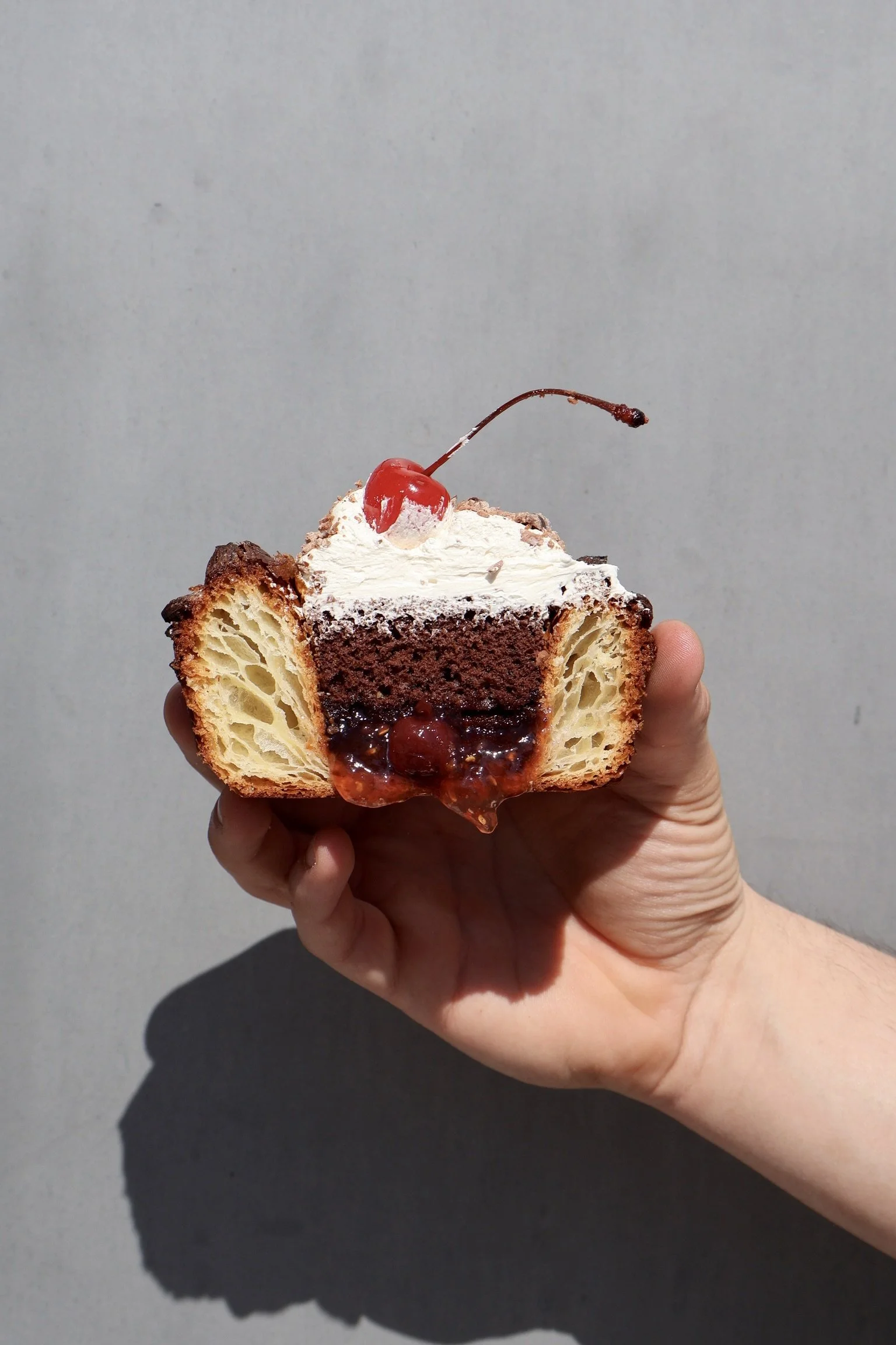 A hand holding a partially sliced cupcake with layers of vanilla cake, chocolate cake, cherry filling, topped with whipped cream and a cherry.