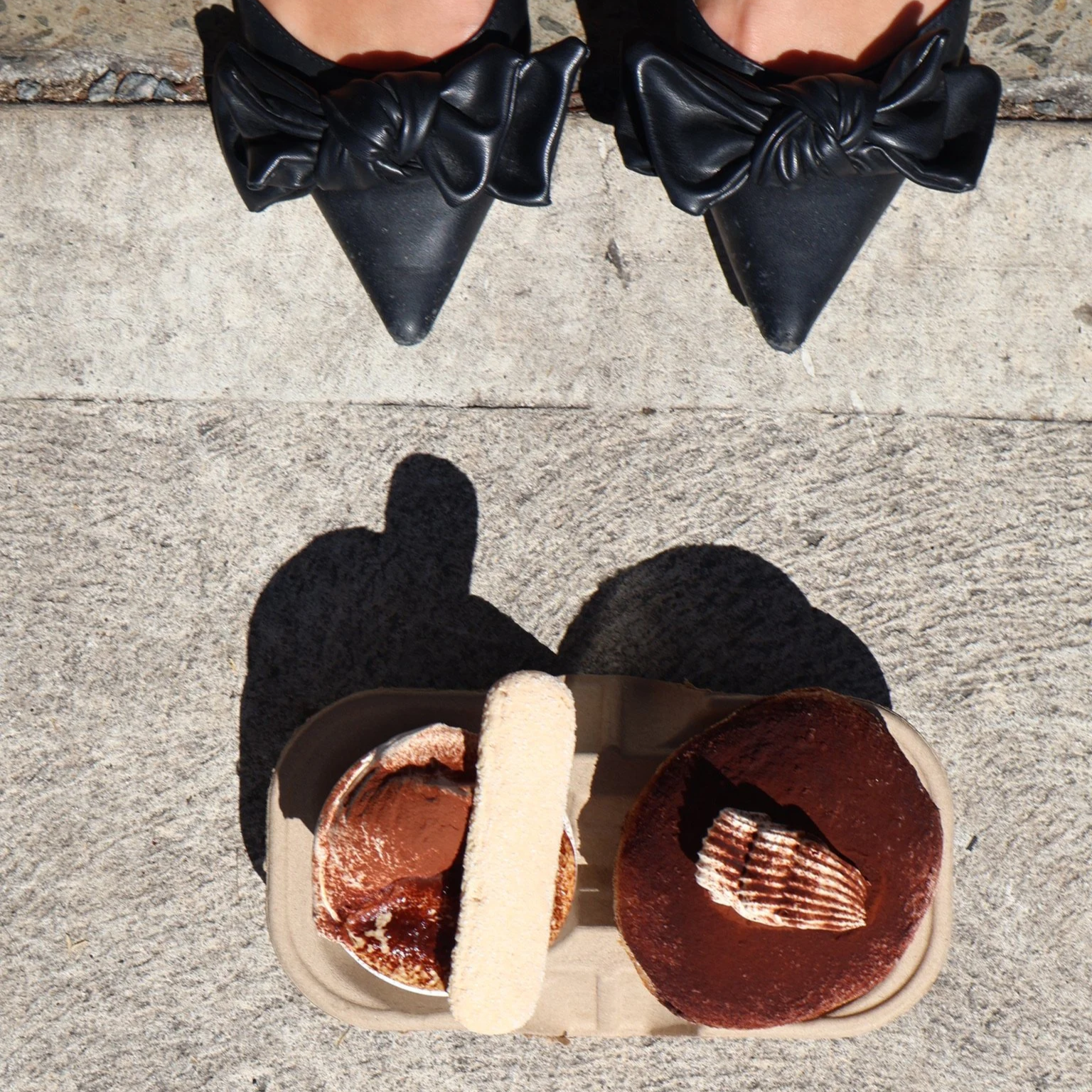 Person wearing black pointed shoes with large bows, standing on concrete sidewalk near a tray of desserts including tiramisu, a chocolate tart, and a white chocolate-covered biscuit.