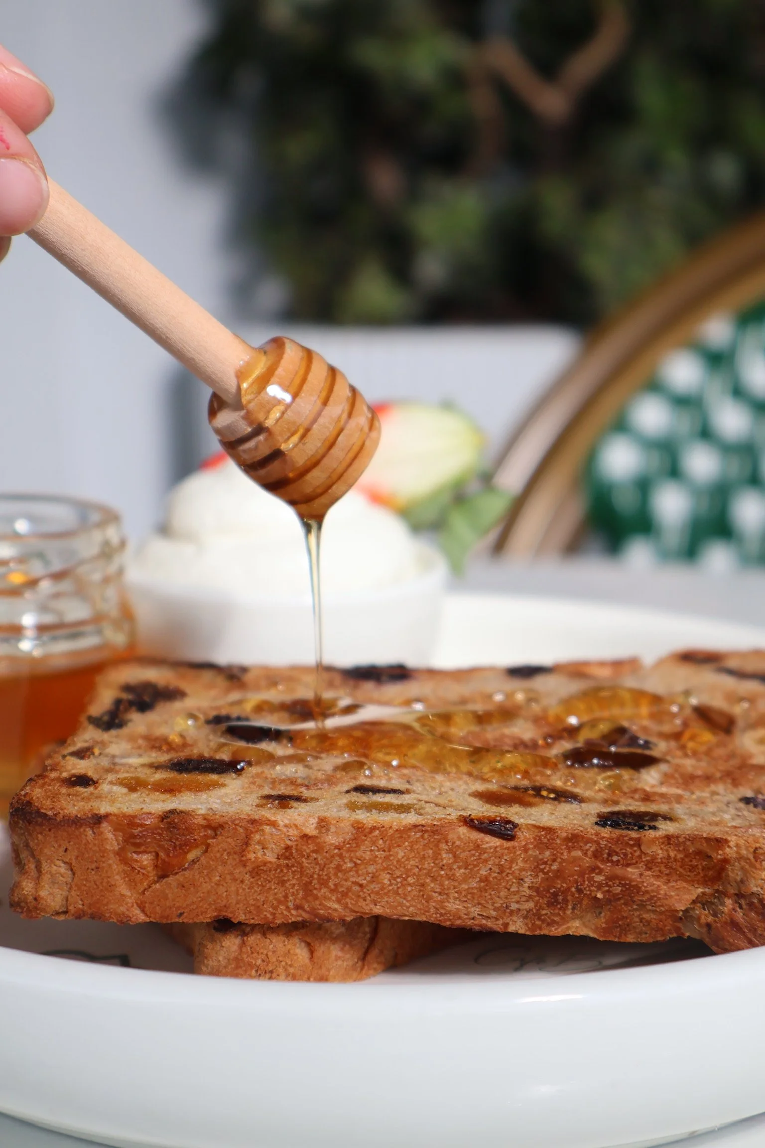 Close-up of a slice of toast with honey dripping onto it, with a jar of honey and a bowl of cream with fruit in the background.