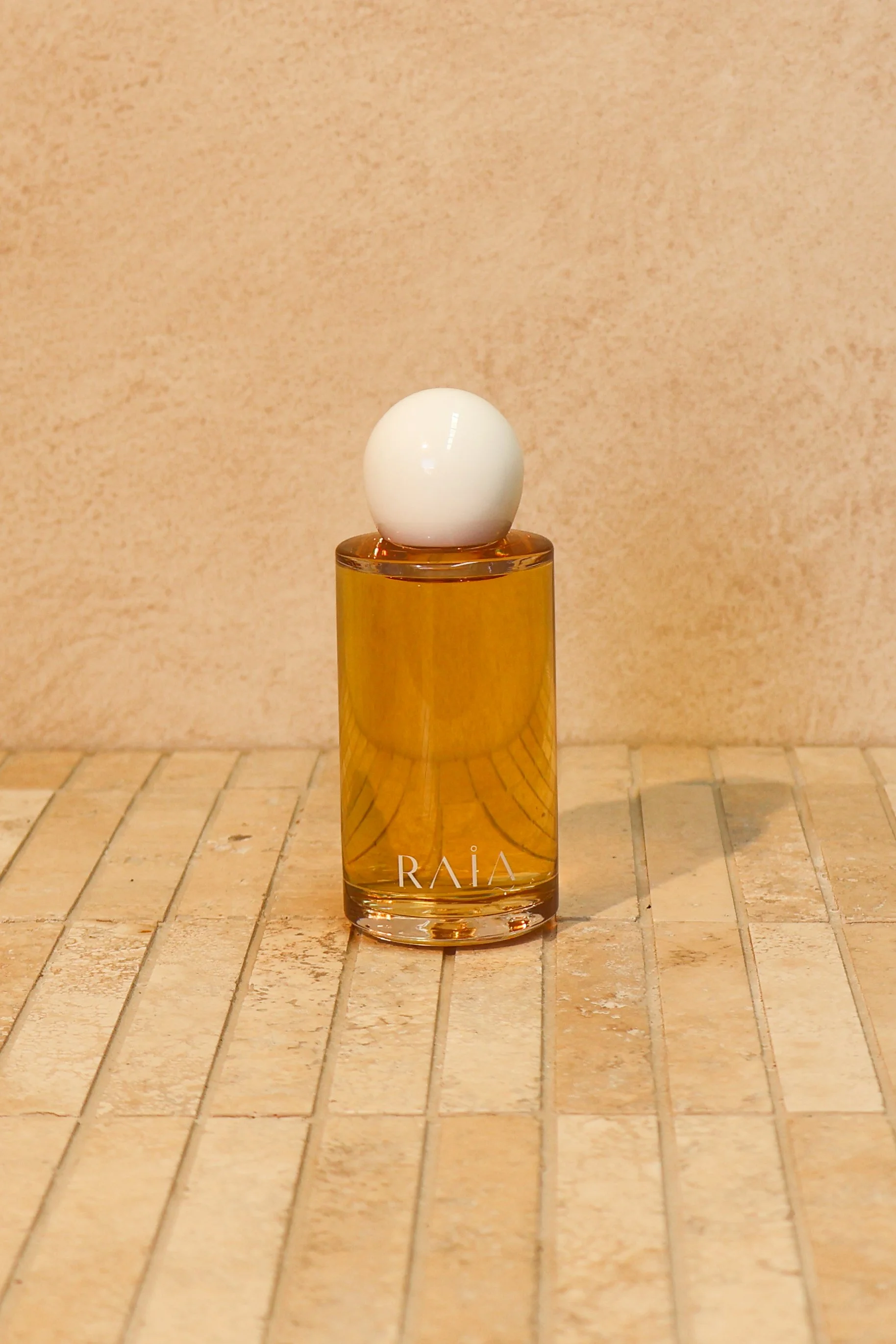 A perfume bottle with a white spherical cap and a transparent amber-colored body, labeled 'RAIA'. It is placed on a tiled surface against a beige wall.