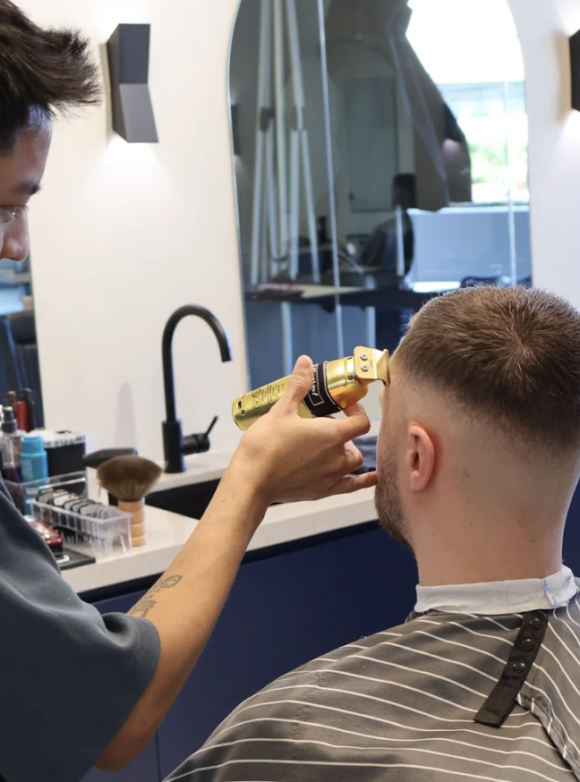 Barber using electric clipper to cut a man's hair in a modern barbershop with a white wall, black faucet, mirror, and various grooming products on the counter.