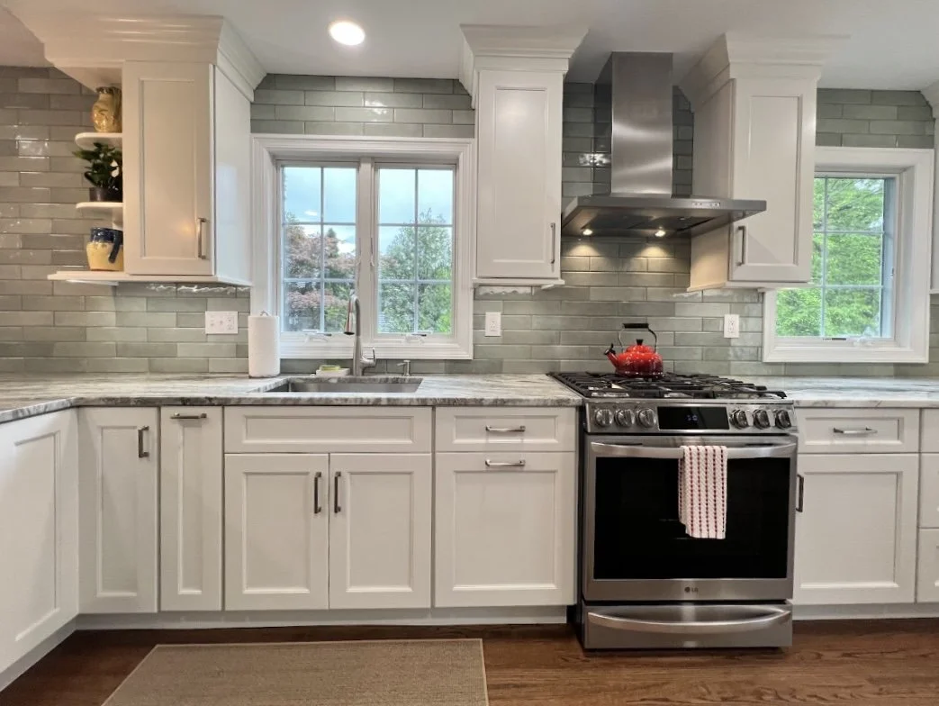 A modern kitchen with white cabinets, a gray tile backsplash, and a stainless steel stove with a red kettle on top. There are two windows above the countertop, allowing natural light to enter.