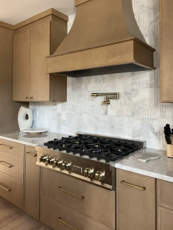 Modern kitchen with beige wooden cabinets, a stainless steel gas stove, a brass range hood, a marble countertop, and a brass wall-mounted faucet. Small black utensils holder on the right, an electric kettle and a plate on the left countertop.