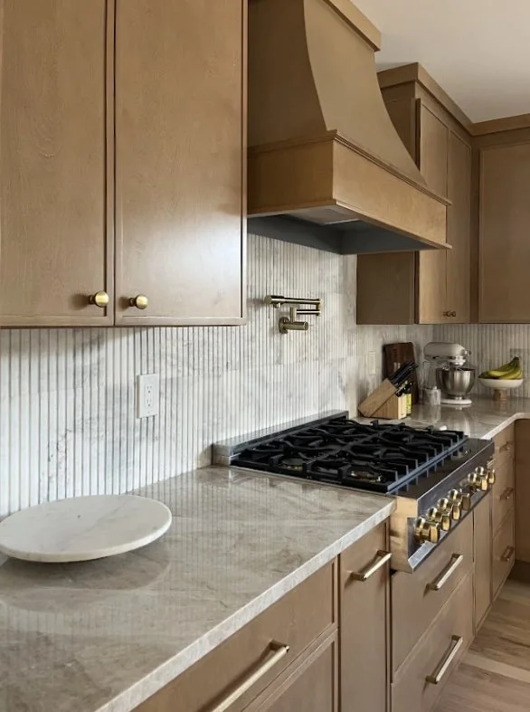 A modern kitchen with light wood cabinets, a beige marble countertop, a gas stove with gold knobs, and a striped tile backsplash. There are kitchen utensils, a bowl of bananas, a stand mixer, and a knife block on the counter.