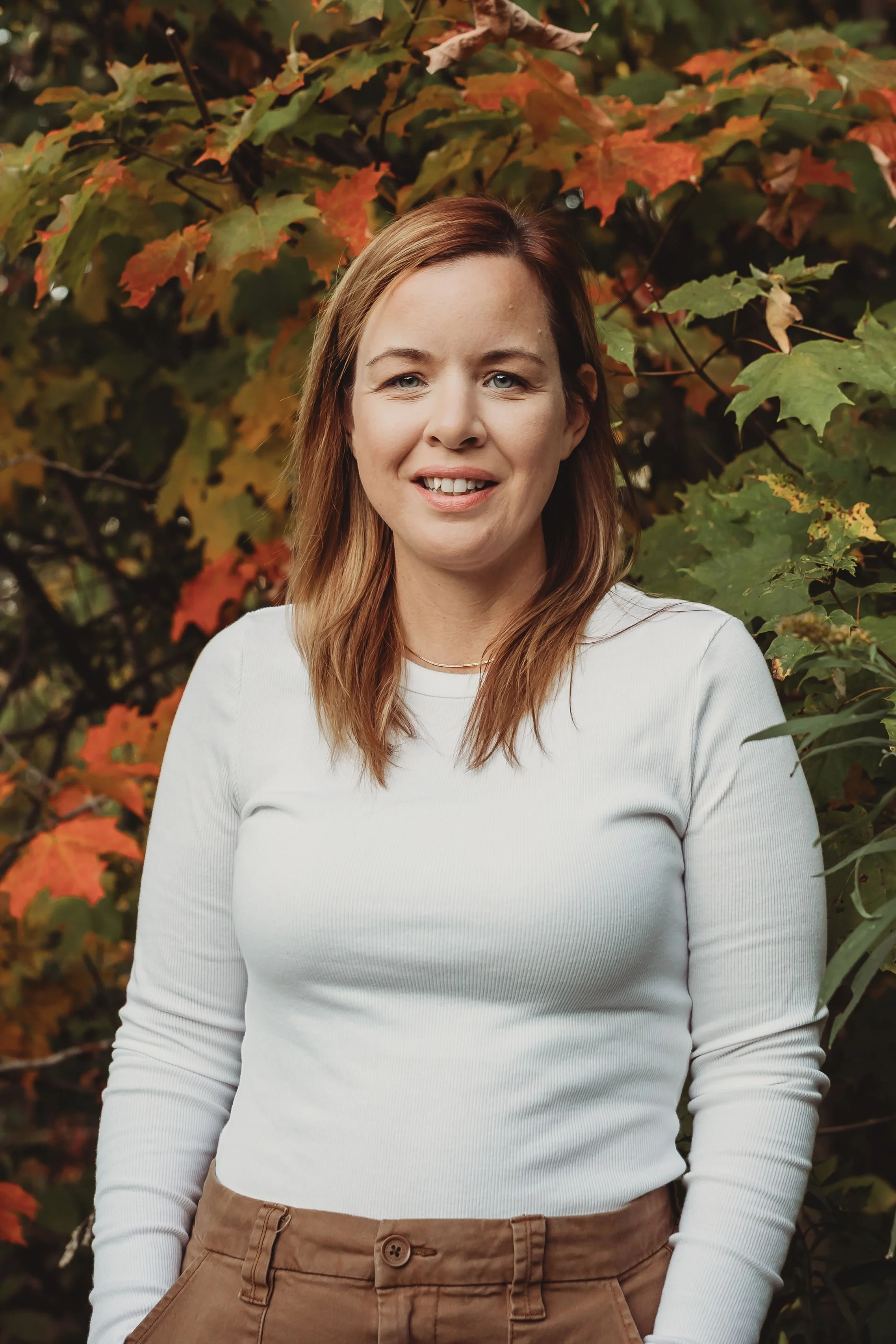 A young woman with shoulder-length light brown hair, wearing a white long-sleeve shirt and tan pants, standing outdoors in front of a background full of colorful autumn leaves.