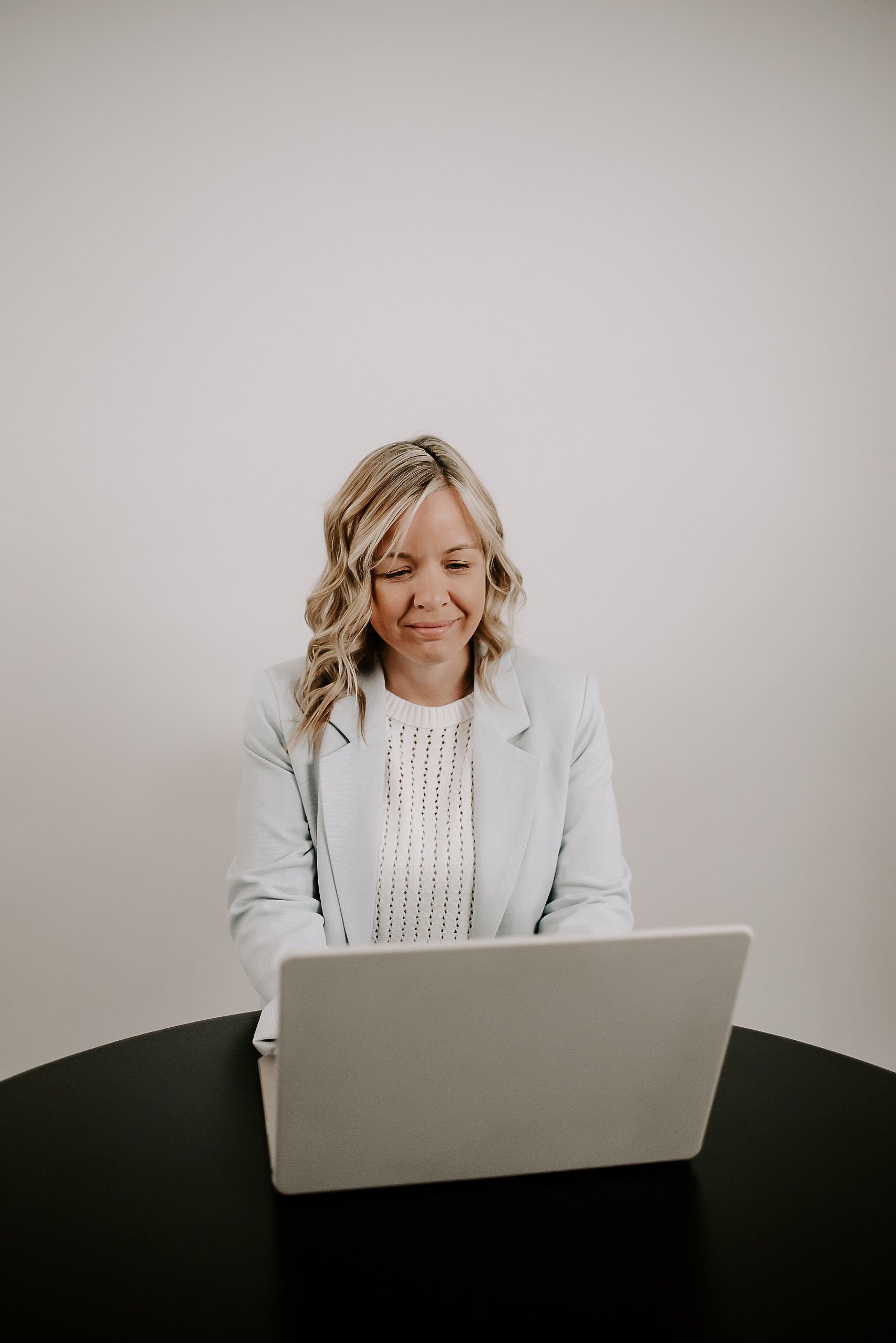 A woman with blonde wavy hair in a light-colored blazer and white top sitting at a black table and looking at a laptop, with a plain white wall in the background.