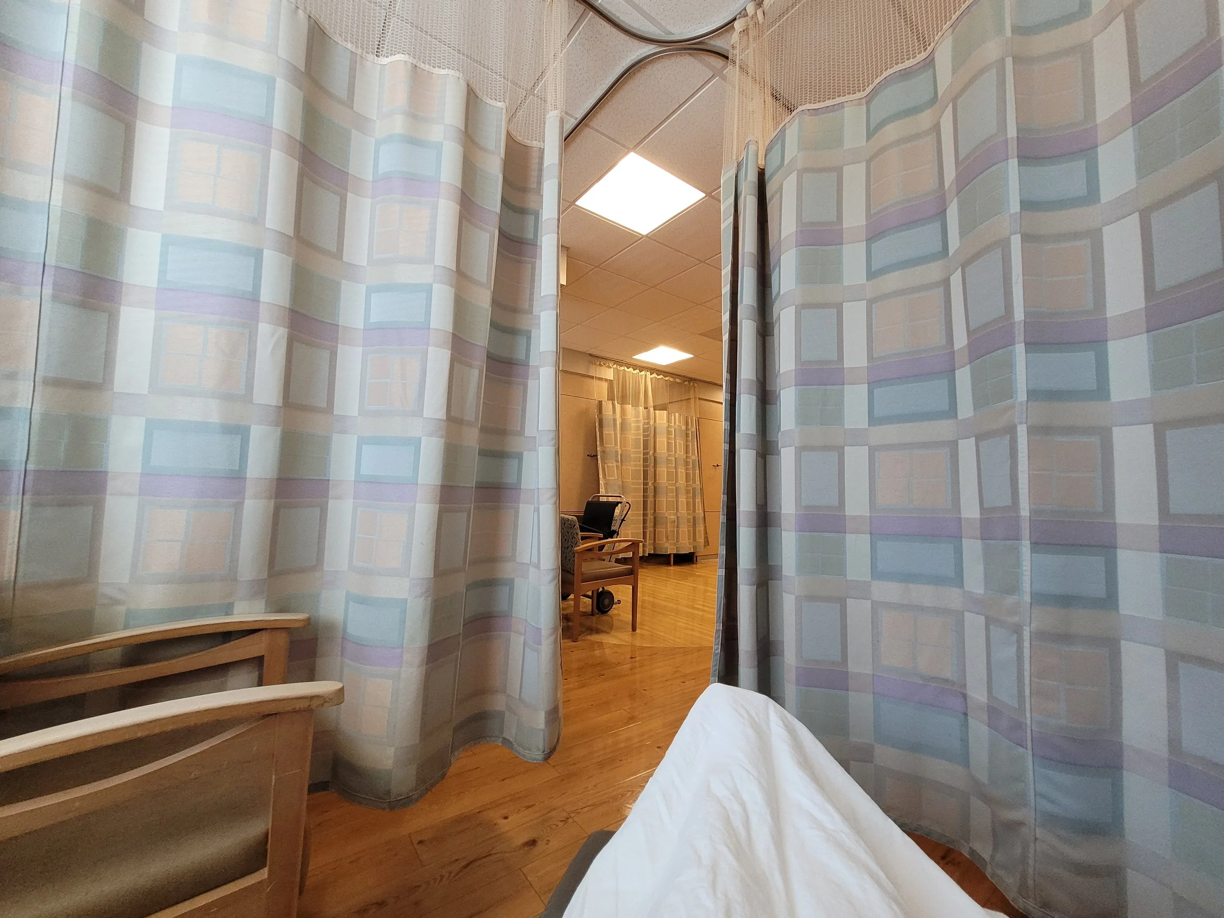Cancer treatment room with partition curtains, hospital recliner with white blanket, chairs, and medical equipment in the background.