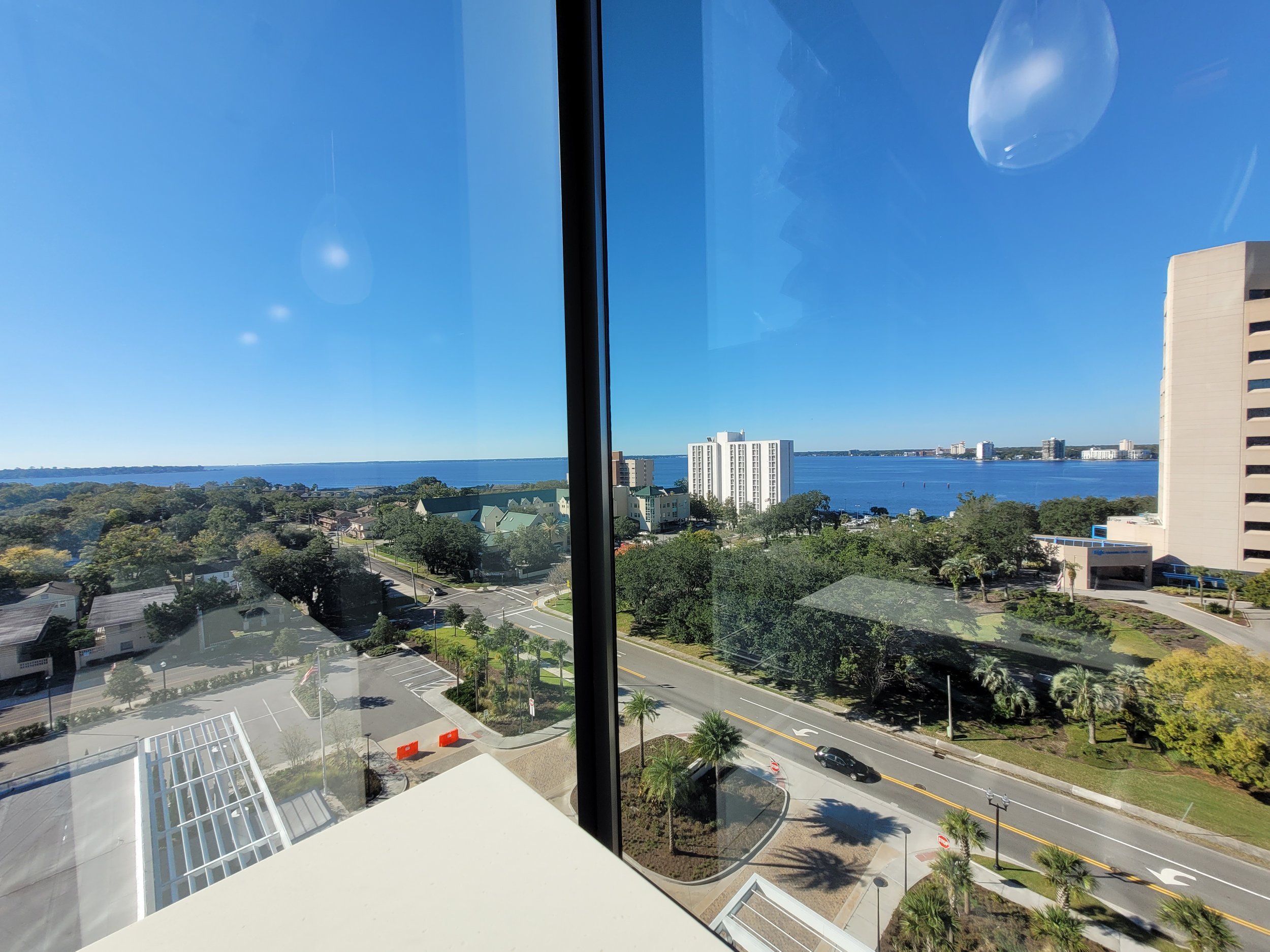 View from a high-rise window showing a cityscape with trees, residential buildings, and a large body of water under a clear blue sky at one of Dana's appointments.