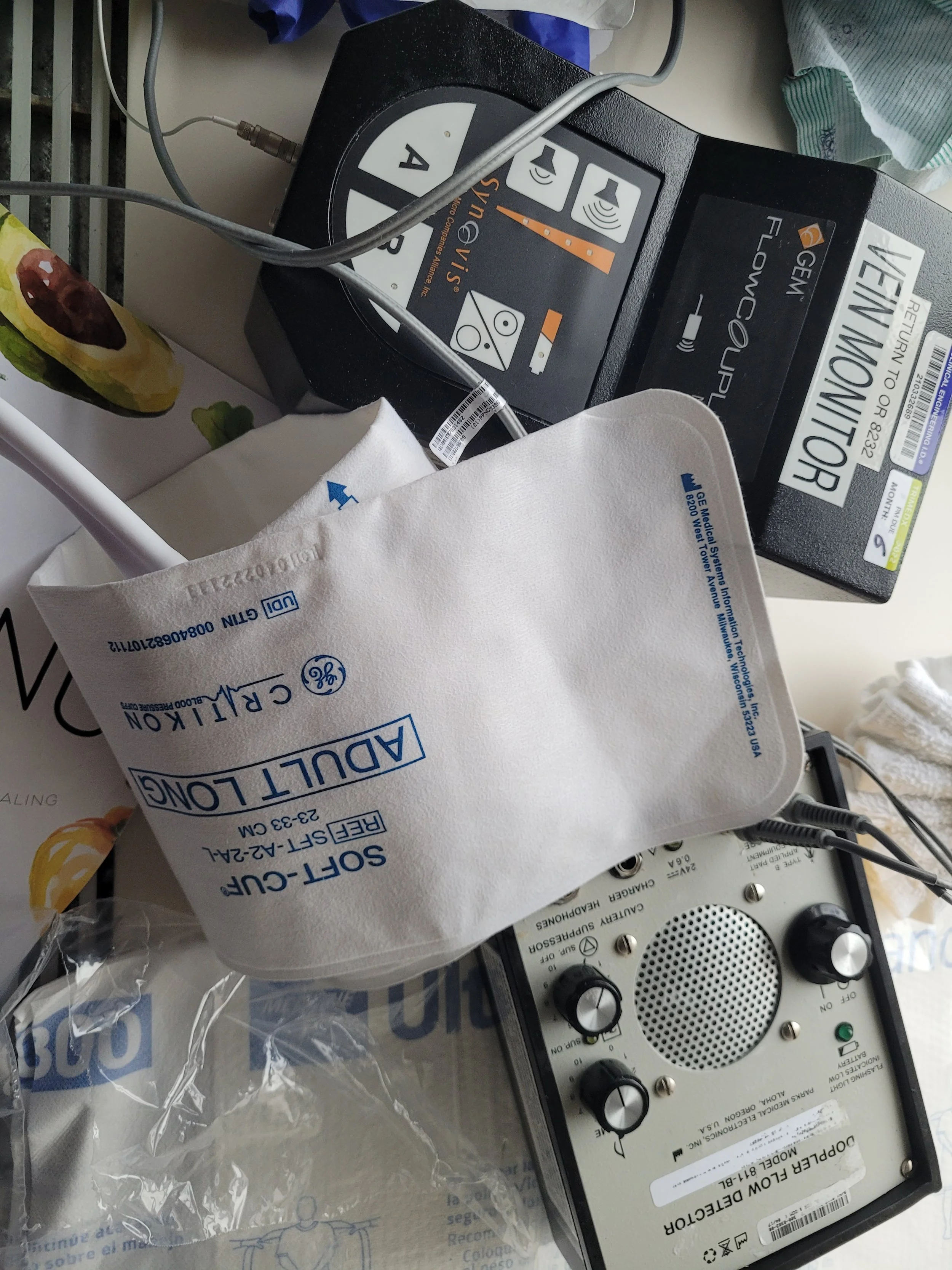 Medical equipment on a table, including a portable ultrasound device, a defibrillator with a paper label, and a small speaker or amplifier. There is a white cloth with blue text covering part of the equipment.