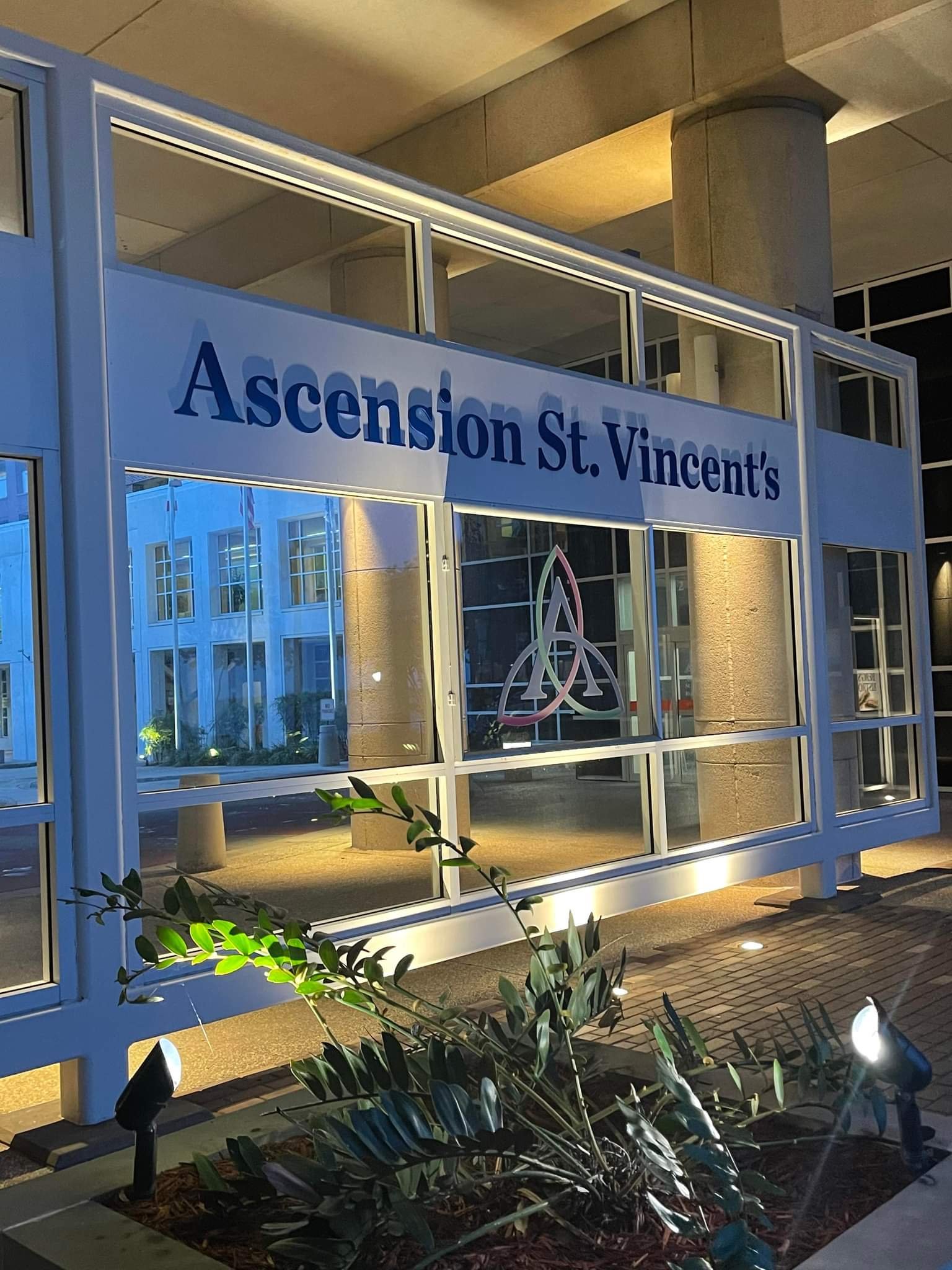 Nighttime view of a modern building with a white framed structure displaying a sign that reads 'Ascension St. Vincent's' and a colorful Christian triquetra symbol behind glass, with some greenery and small lights in the foreground.