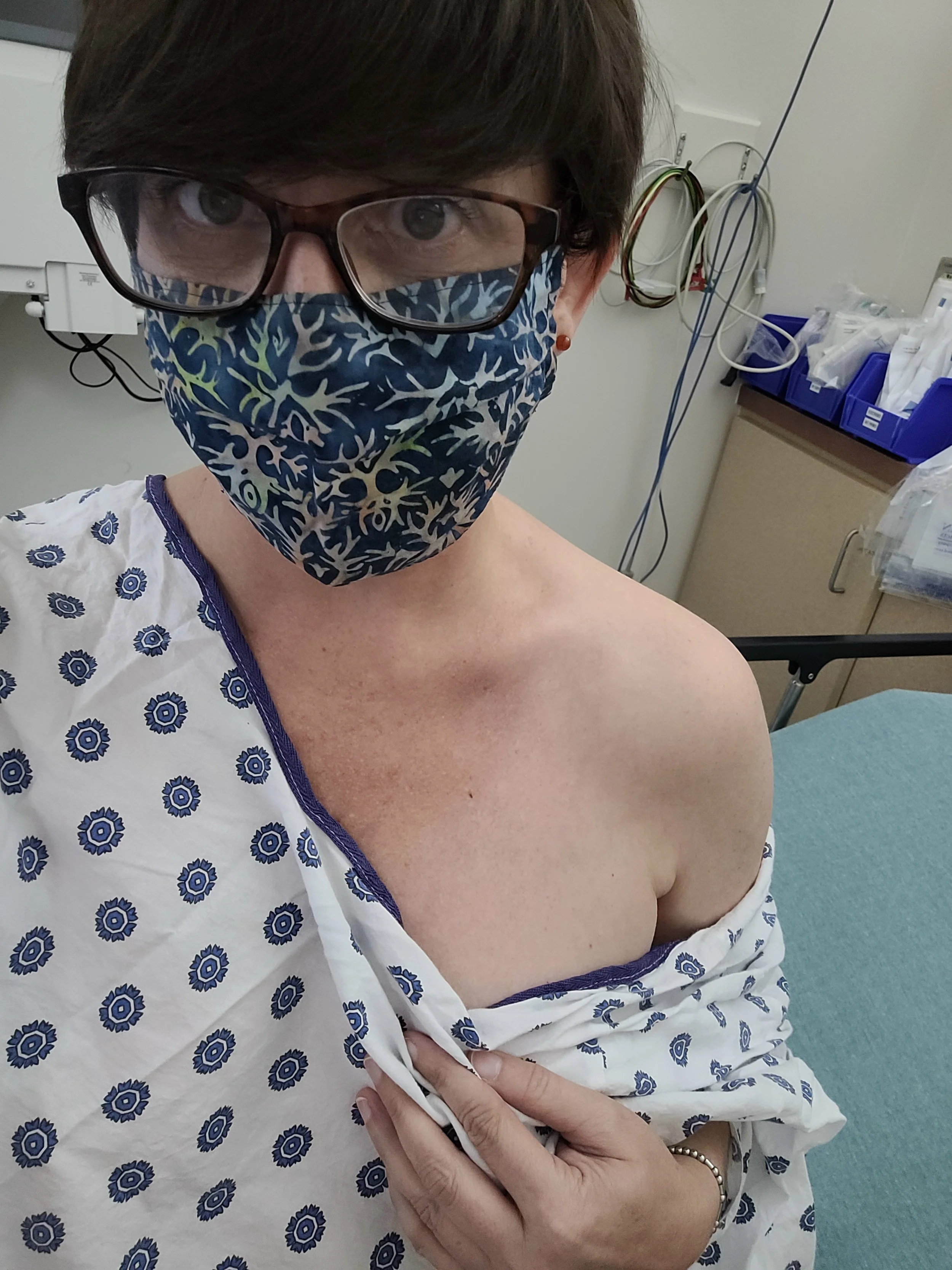 Dana in a hospital gown and face mask taking a selfie in a medical examination room for pre-surgery, with hospital equipment and supplies visible in the background.