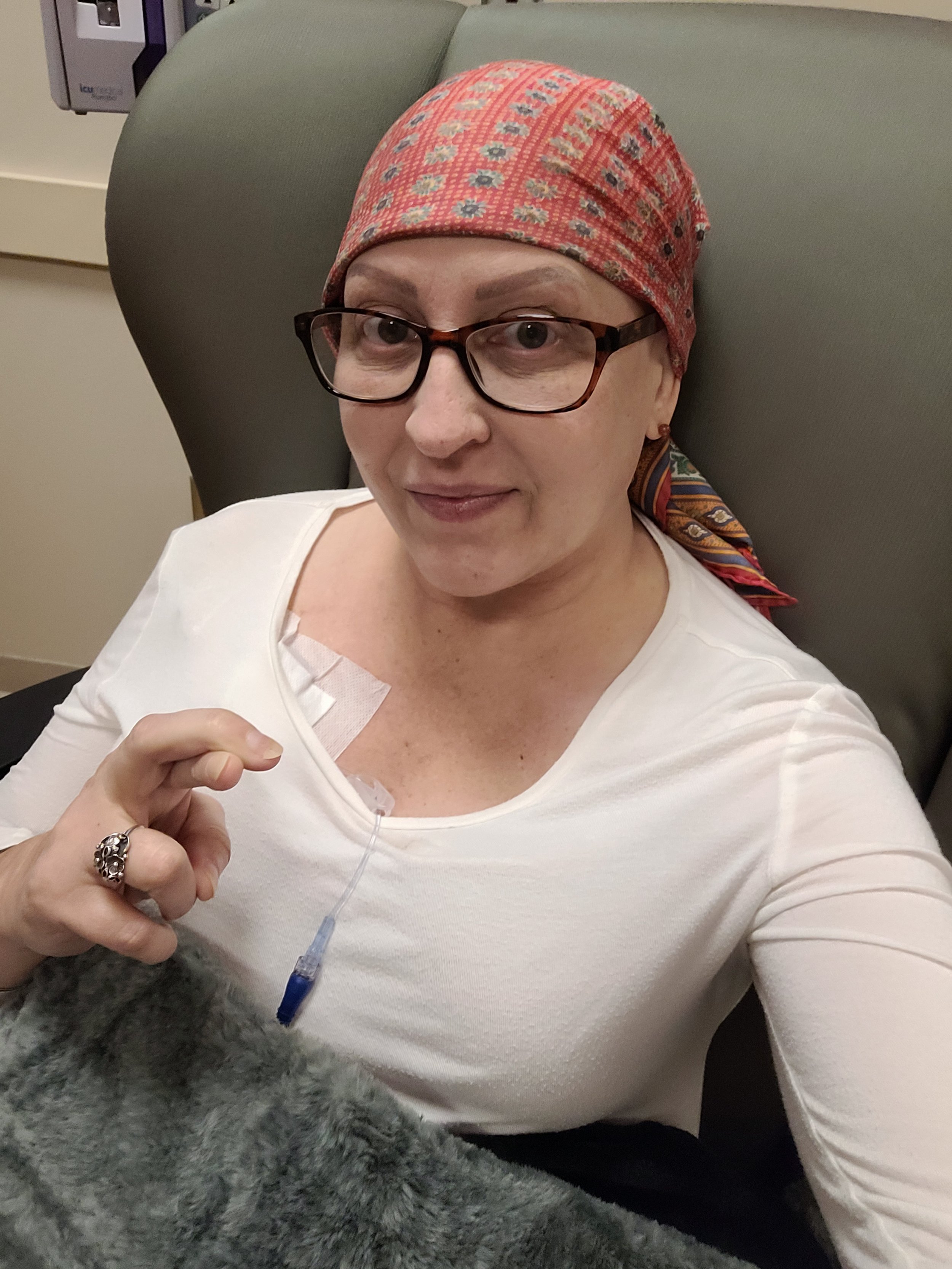 Dana Lupo at cancer center getting chemotherapy with a bandana, glasses, and a white shirt. She has an IV and is making a fingers crossed gesture. She is smiling at the camera.