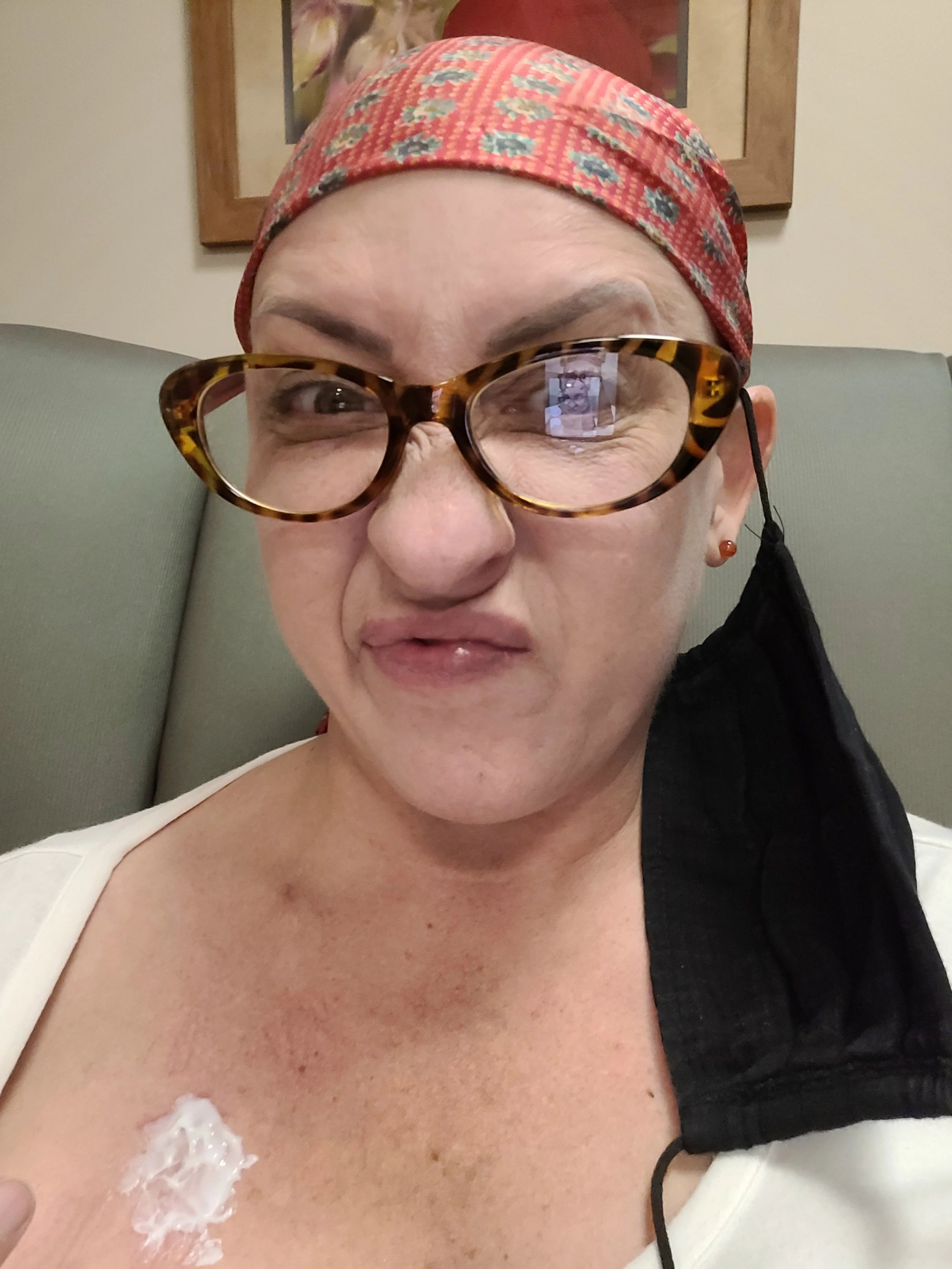 Dana with glasses, a red patterned headscarf, and a face mask hanging off one ear, making a grimace. She has a bandage and some redness on their chest and are seated against a plain background with a framed picture.