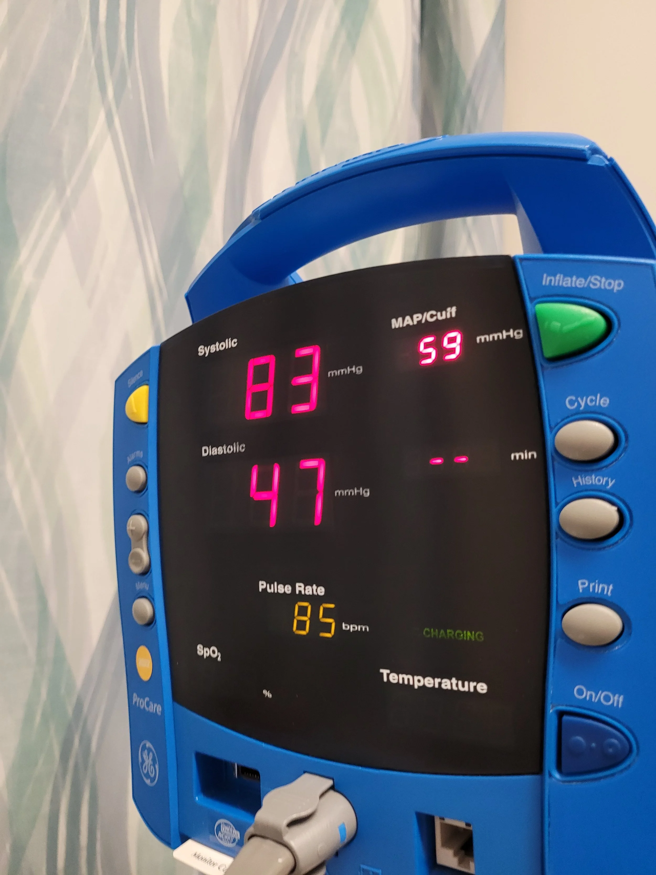 A digital blood pressure monitor displaying a systolic reading of 83 mmHg, a diastolic reading of 47 mmHg, and a pulse rate of 85 beats per minute, with a blue casing and control buttons on the side.