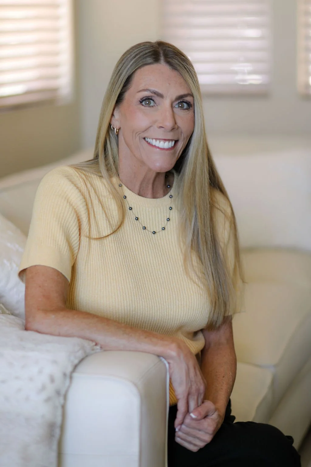 A smiling woman with long blonde hair sitting on a beige couch in a bright room with window blinds.