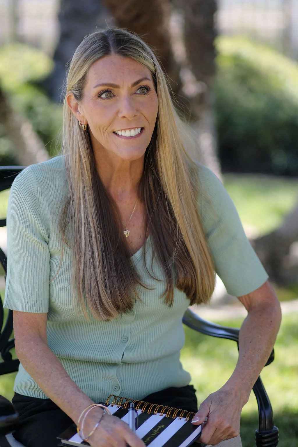 Smiling woman sitting outdoors on a bench, holding a striped planner, with trees and greenery in the background.