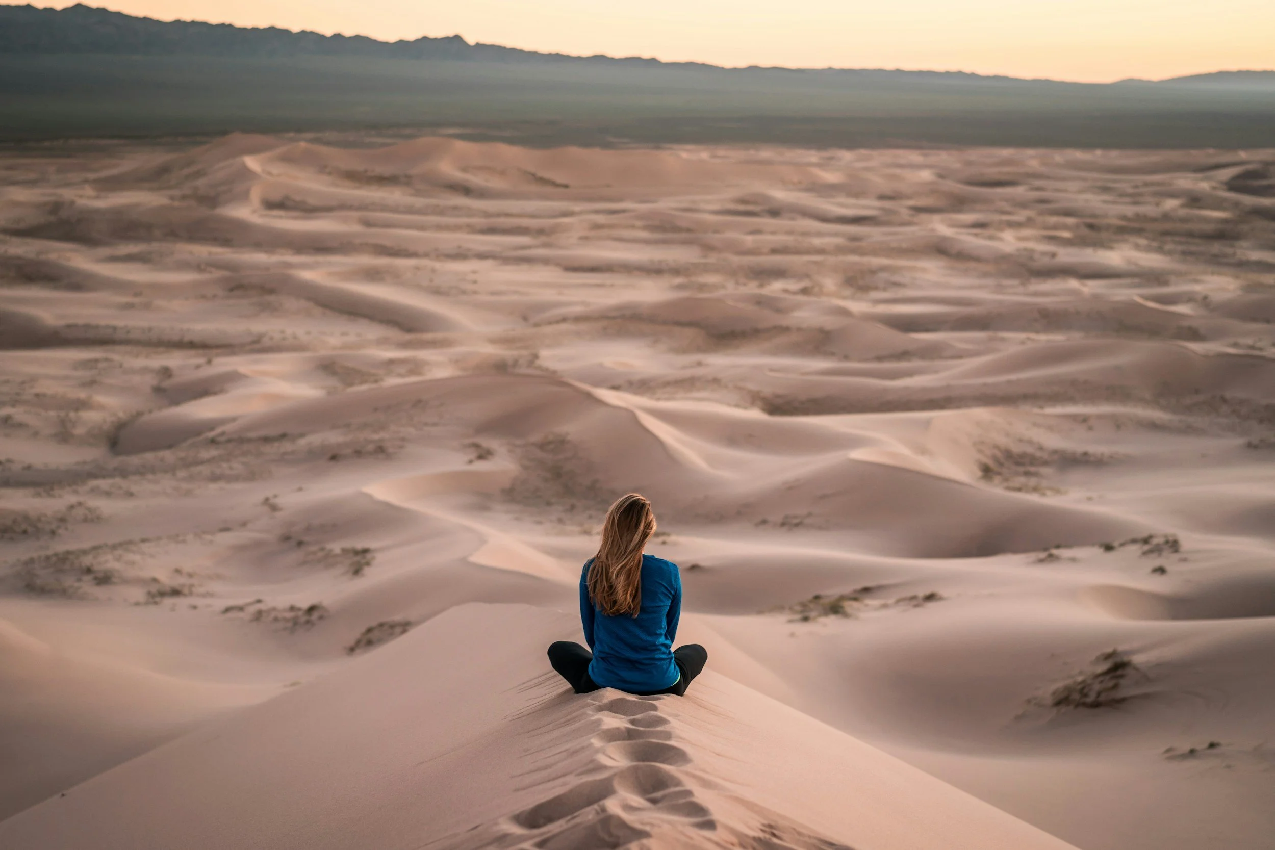 A woman in a blue jacket sitting cross-legged on a sand dune in a desert, facing away, with footprints leading to her, overlooking expansive sand dunes at sunset.