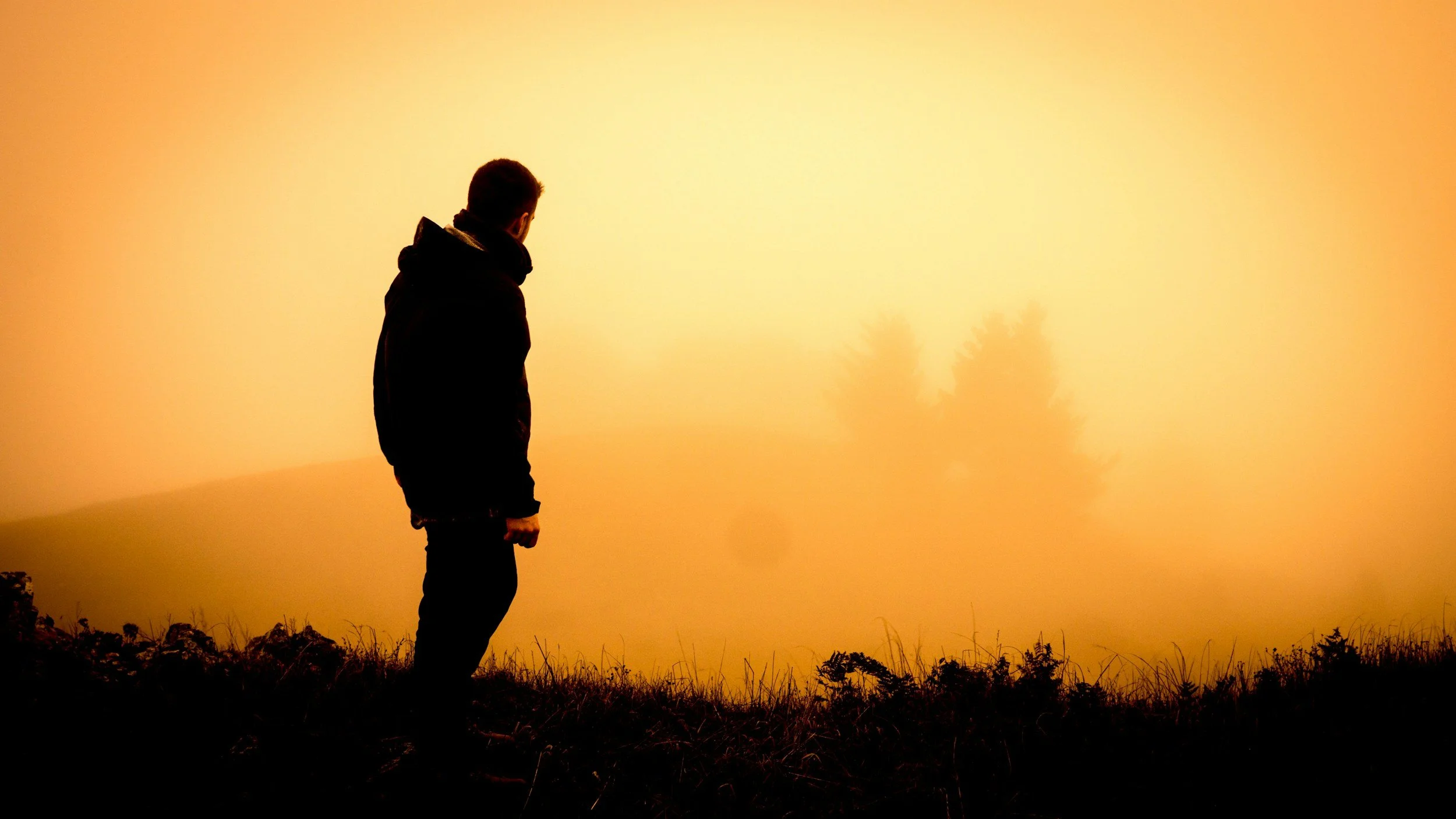 Silhouette of a man standing outdoors at sunset or sunrise, with a hazy sky and faint trees in the background.