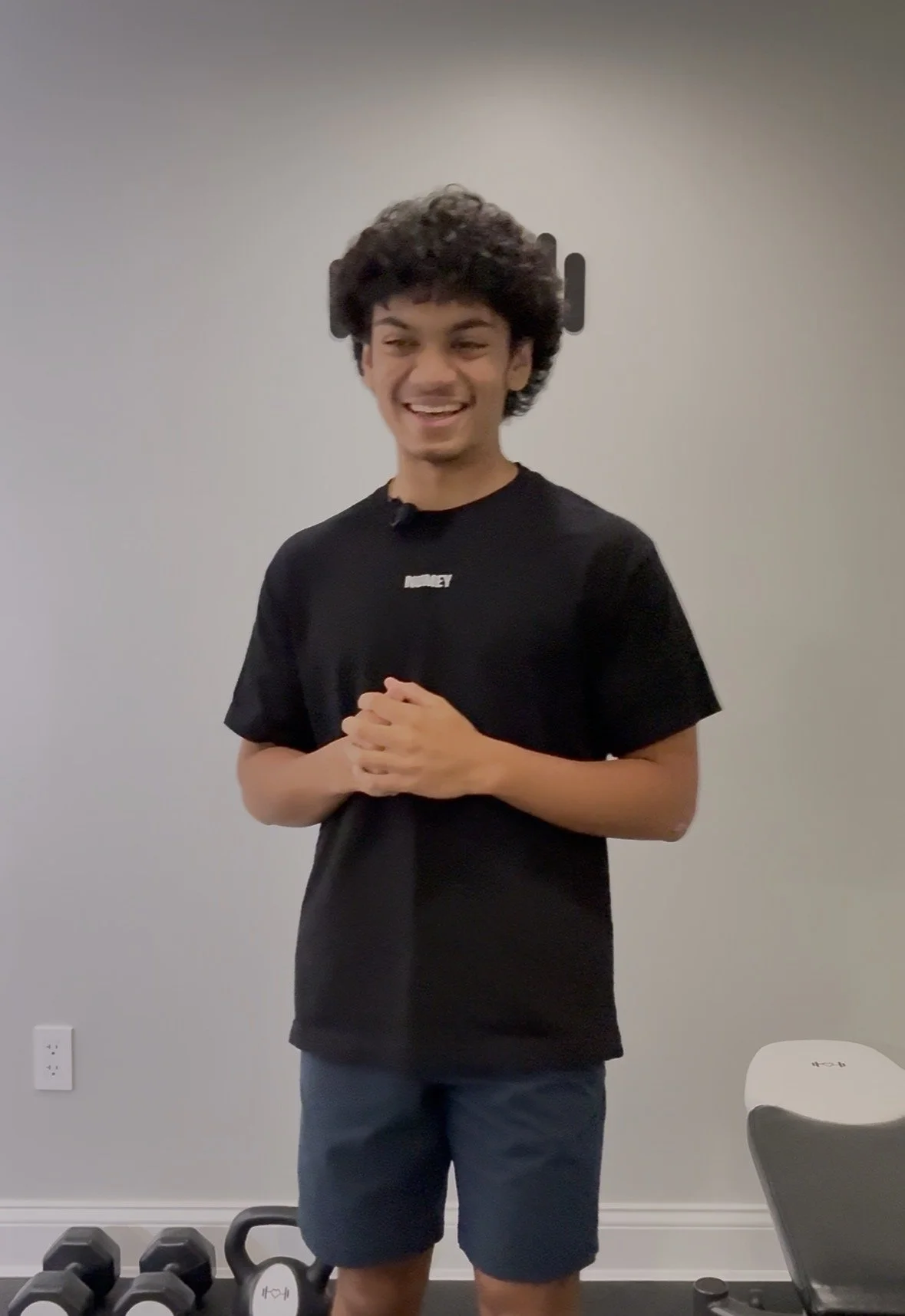 A young man with dark, curly hair smiling and standing in a home gym with dumbbells and a kettlebell on the floor.
