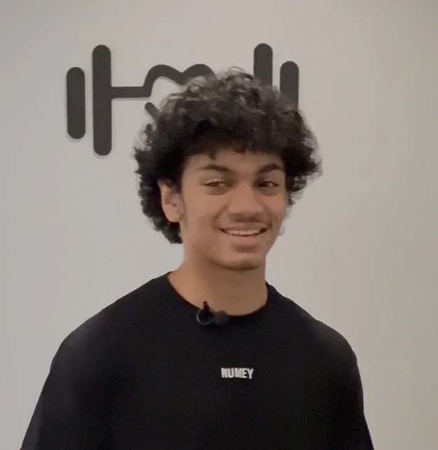 A smiling woman with shoulder-length black hair wearing a blue top, standing in front of green plants.