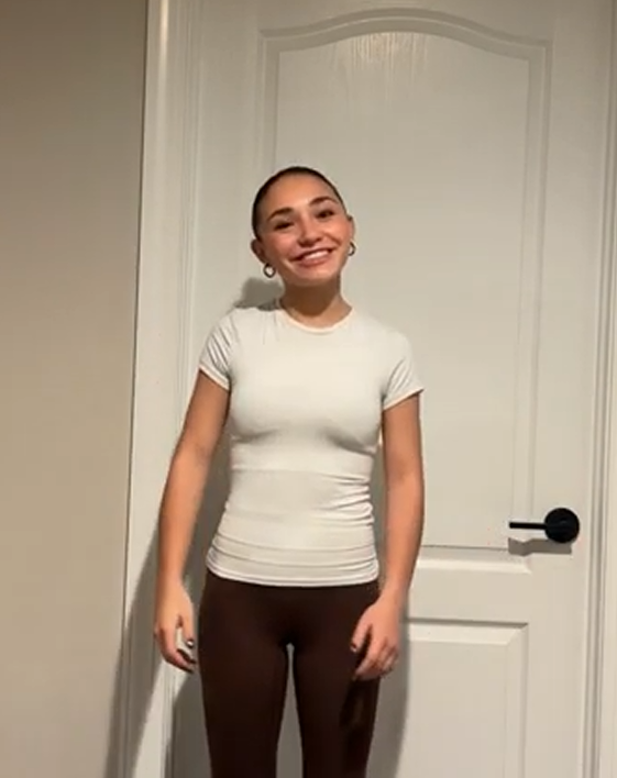 A smiling young woman with brown hair pulled back, wearing a white fitted t-shirt and dark pants, standing in front of a closed white door in a room with beige walls.