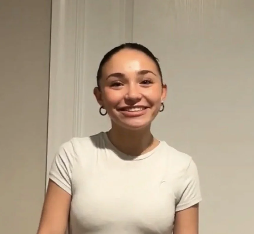 Smiling woman with glasses, hoop earrings, and necklace, wearing a striped shirt, indoors with blurred background.