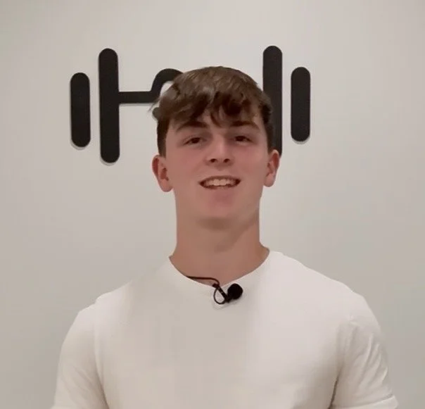Young man with brown hair smiling in front of a white wall with a black fitness logo and dumbbell graphics.