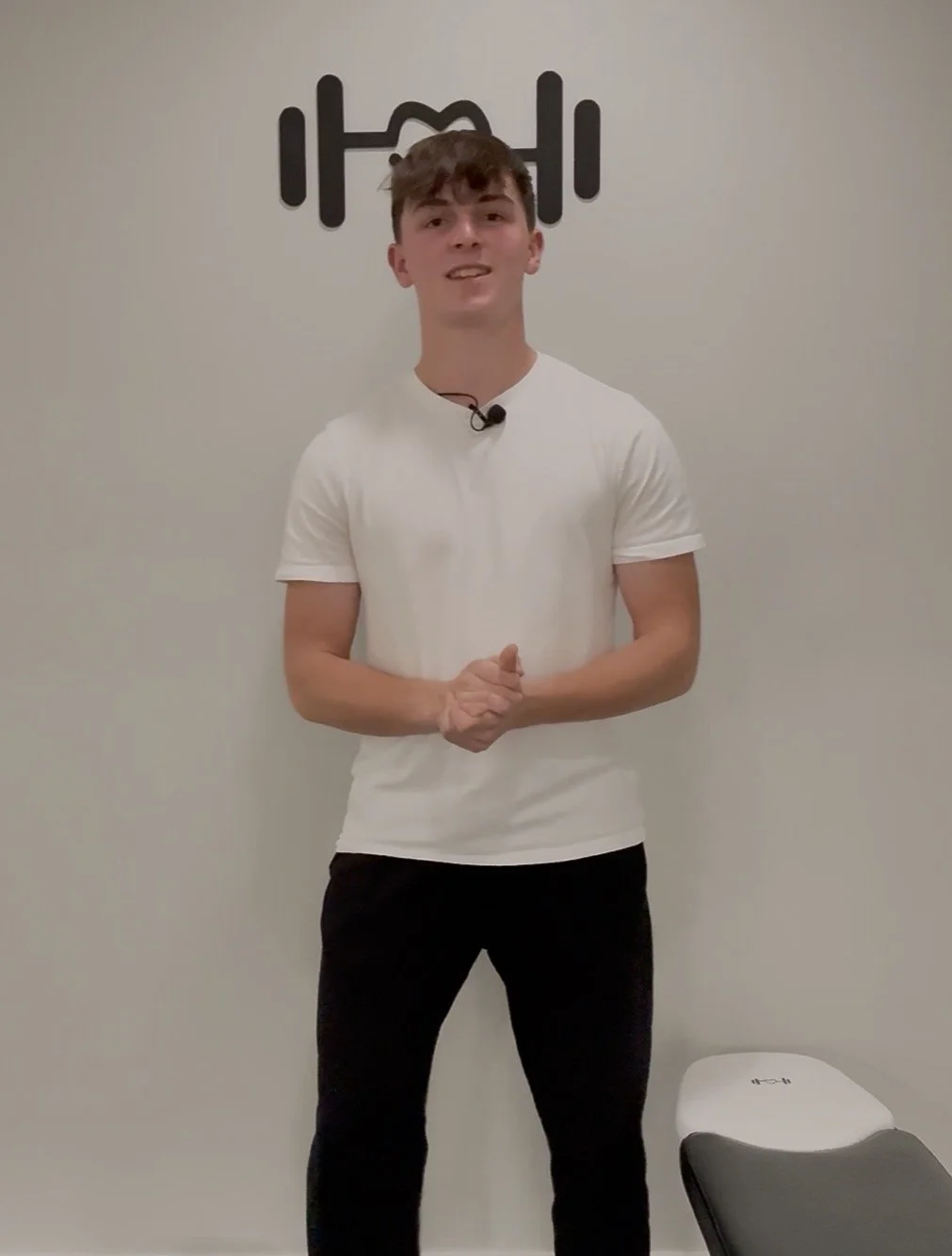 A young man with brown hair, wearing a white t-shirt and black pants, standing in front of a white wall with a black logo of a person lifting weights. There is a white and black weight bench to his right.