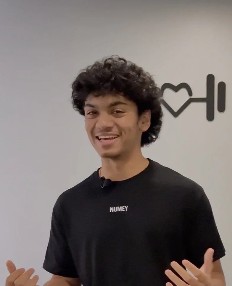 A smiling woman with shoulder-length black hair wearing a blue top, standing in front of green plants.